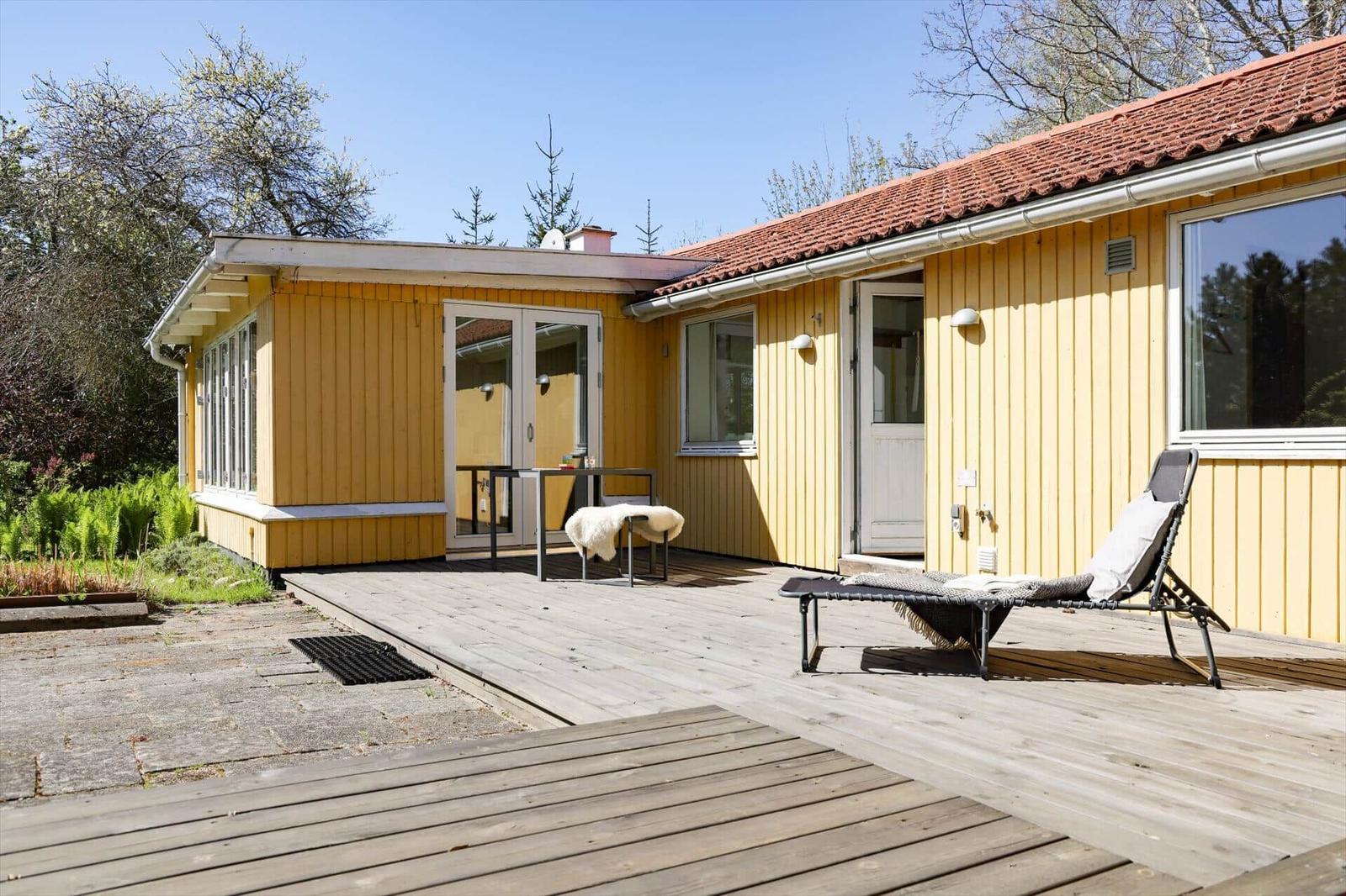 Ein gelbes Haus mit Terrasse, Liegestuhl und Garten. Hintergrund: Bäume und klarer Himmel.