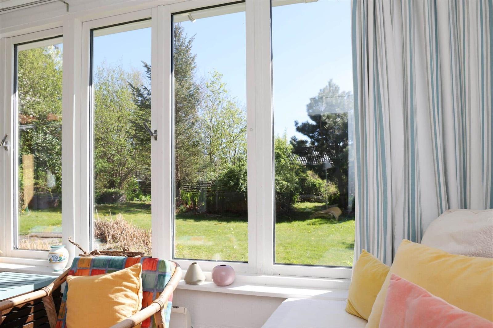 Grosses Fenster mit Blick auf einen grünen Garten. Vor dem Fenster stehen ein Sessel und ein Sofa mit Kissen.
