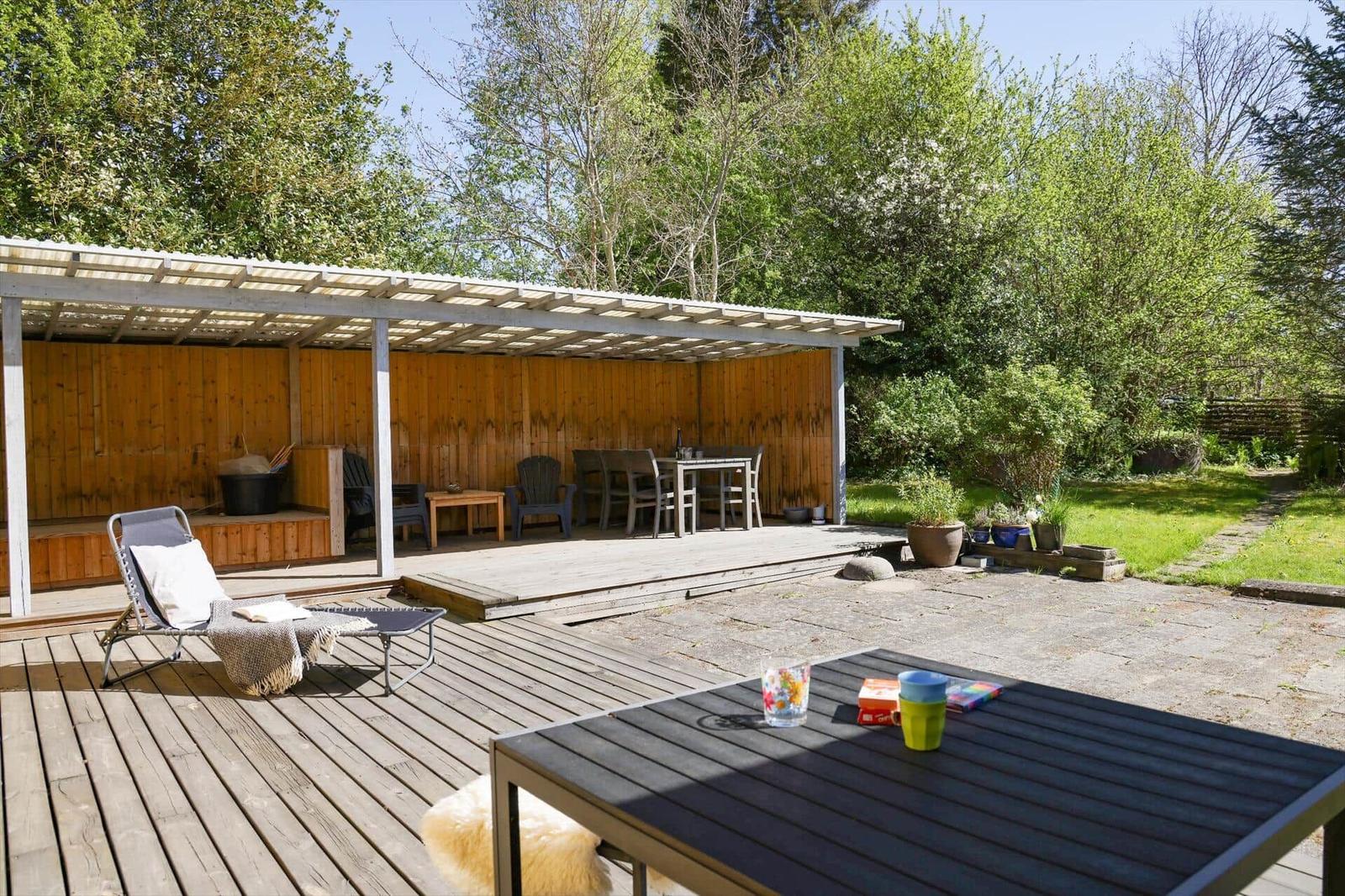 Holzterrasse mit Lounge, Tisch und Stühlen unter Dach. Grün umgeben.