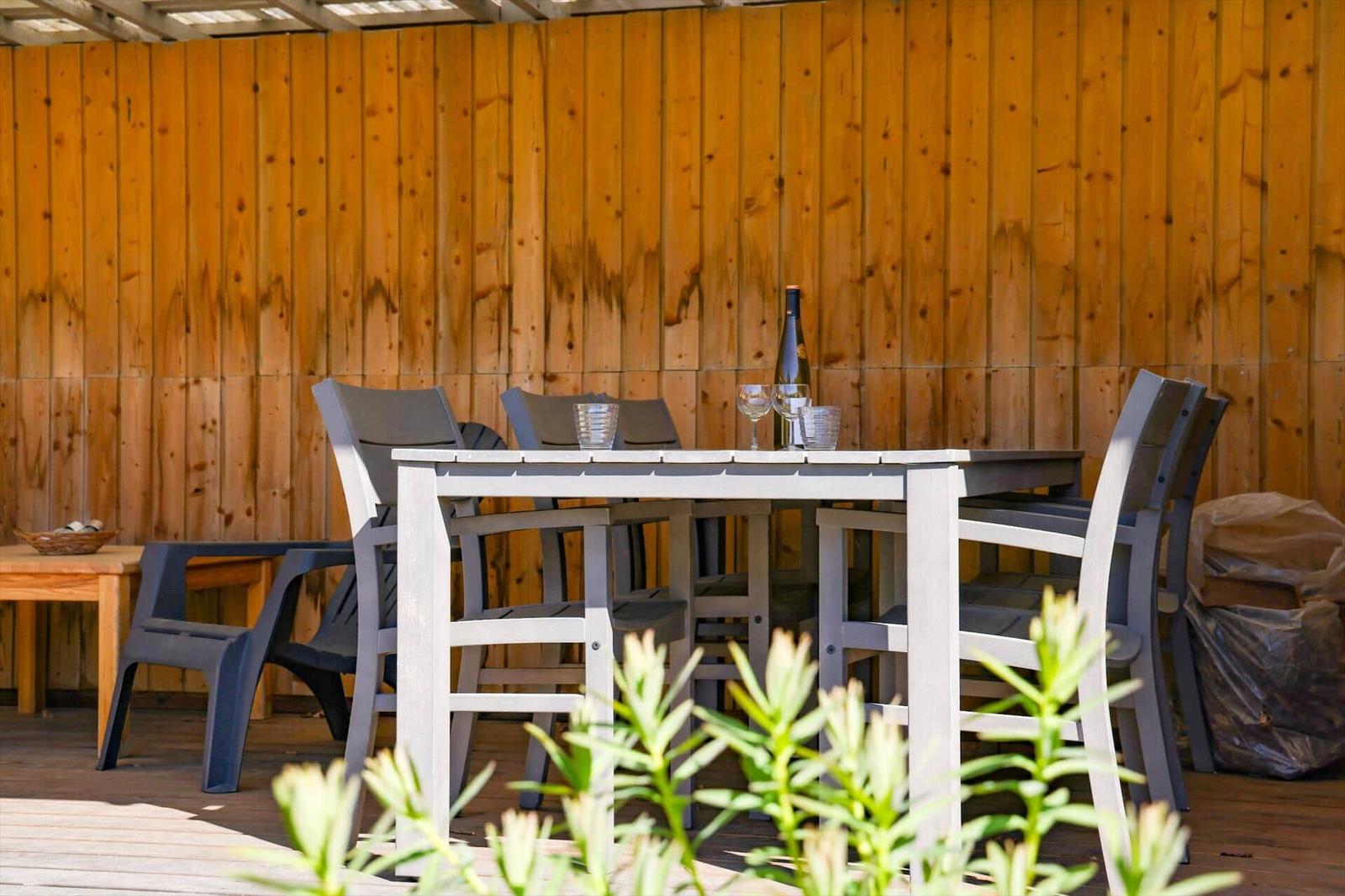 Terrasse mit Tisch und Stühlen, Holzwand im Hintergrund, Pflanzen im Vordergrund.