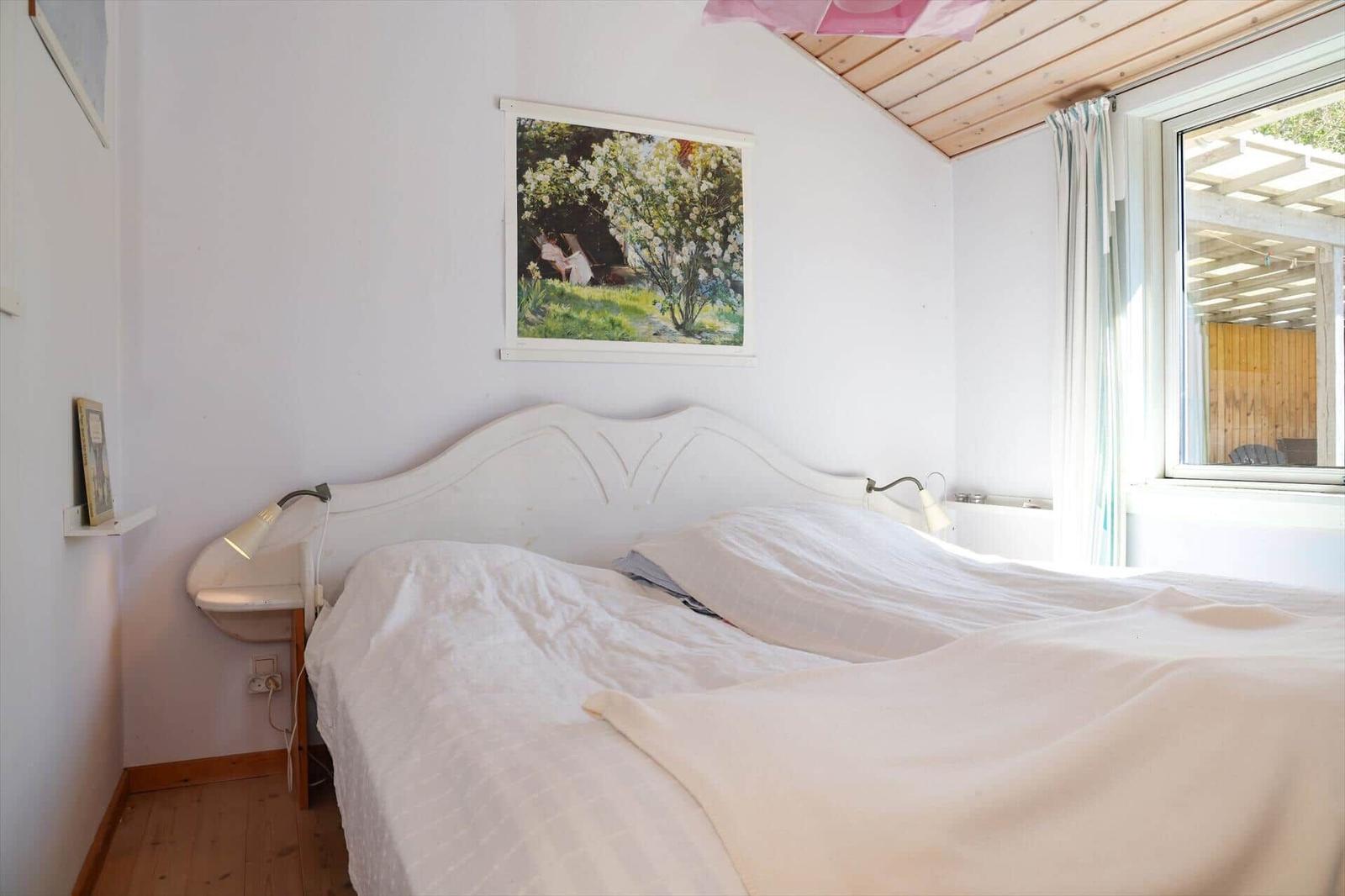Schlafzimmer mit weißem Bett, Wandbild und Fenster mit Blick auf Terrasse.