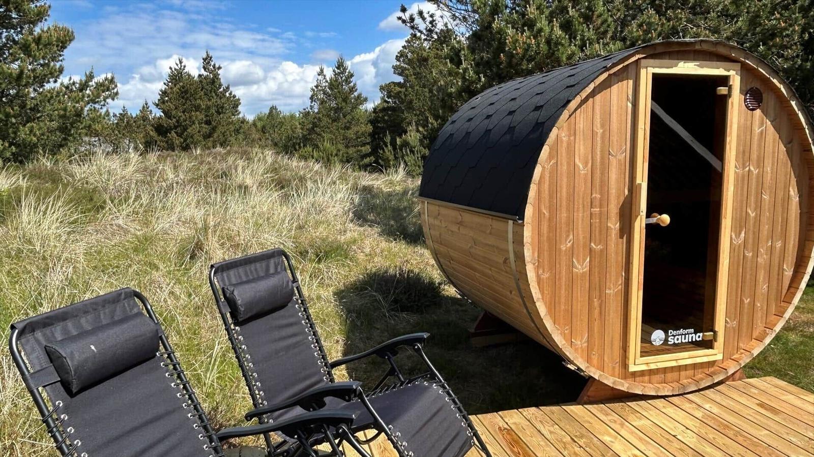 Holzsauna mit offenem Eingang auf Holzterrasse, zwei schwarze Liegestühle im Gras, Hintergrund mit Bäumen.