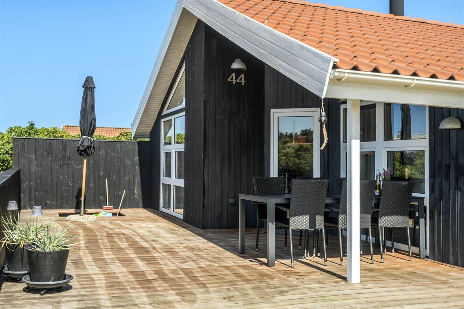 Terrasse mit Tisch, Stühlen und Sonnenschirm. Schwarze Holzwand und roter Dachziegel.