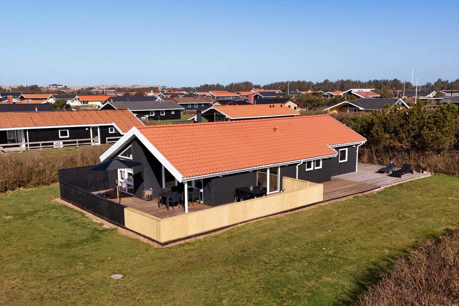 Schwarzes Ferienhaus mit rotem Dach und Holzterrasse. Grün umgeben, Blick auf andere Häuser.