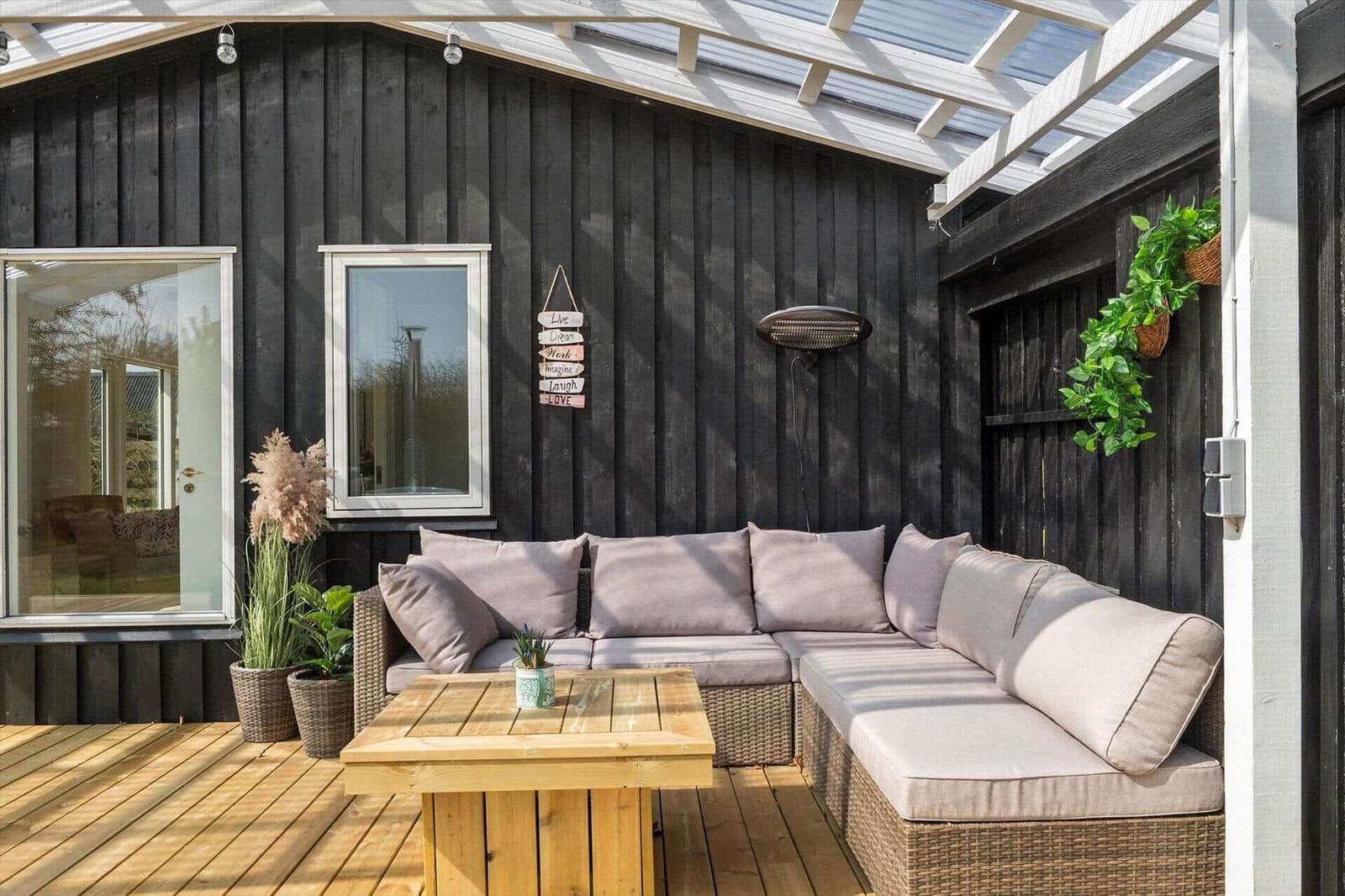 Terrace with seating group, wooden table, and plants against black wood wall under glass roof.