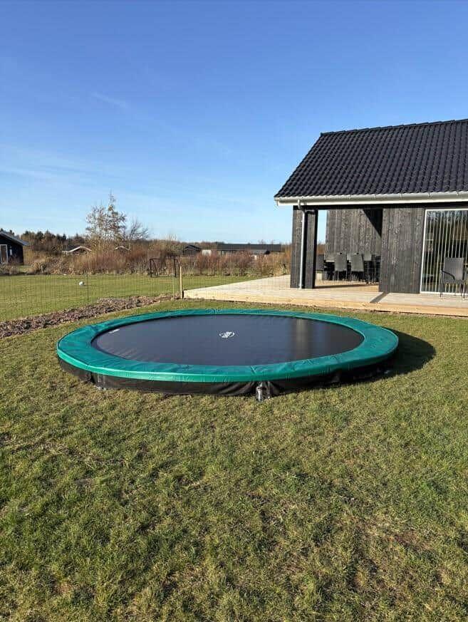 Rundes Trampolin auf grünem Rasen vor einem Holzhaus mit Terrasse.