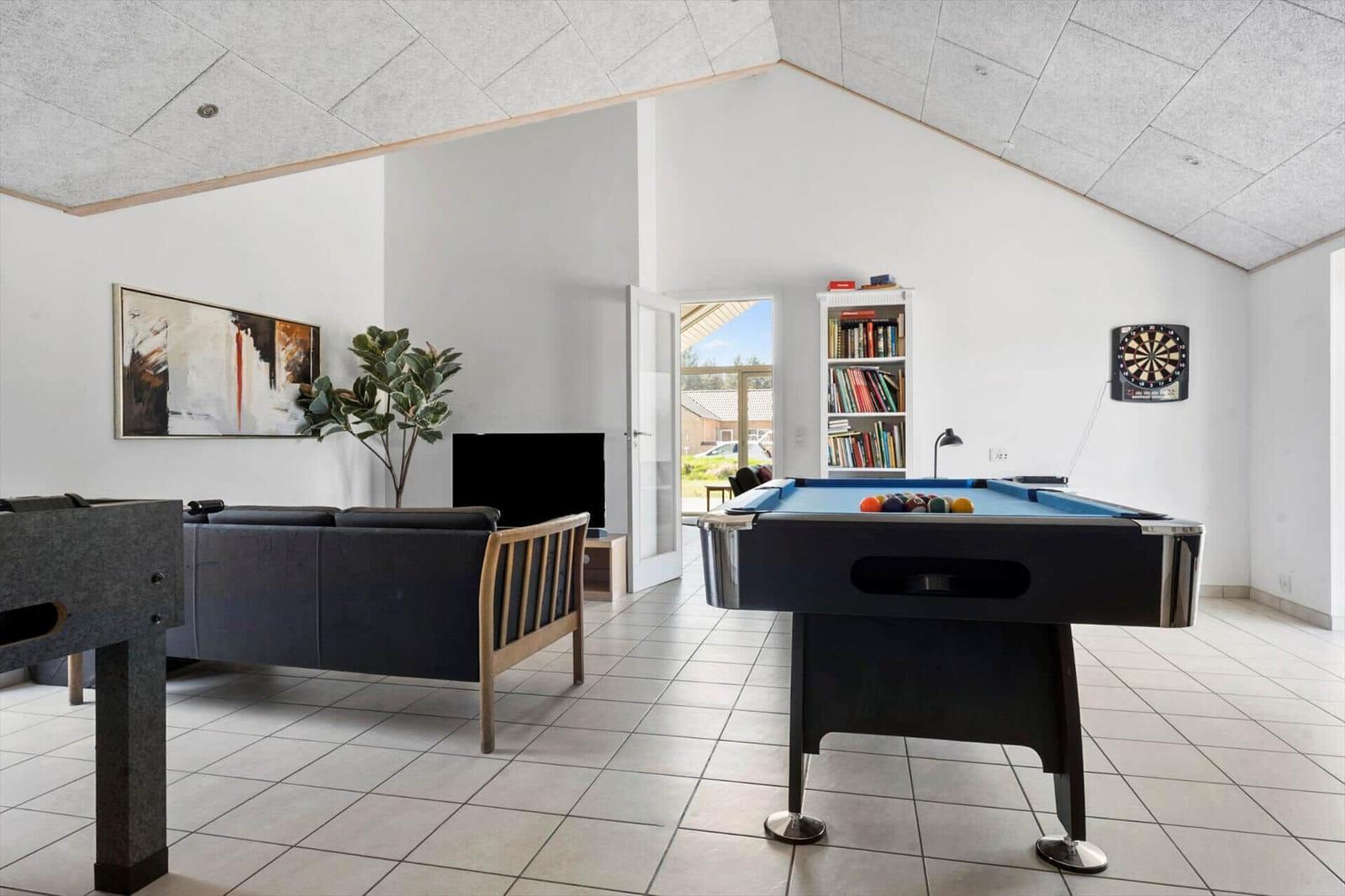 Raum mit Billardtisch, Dartboard und Bücherregal. Couch, Fernseher und Pflanze sichtbar.