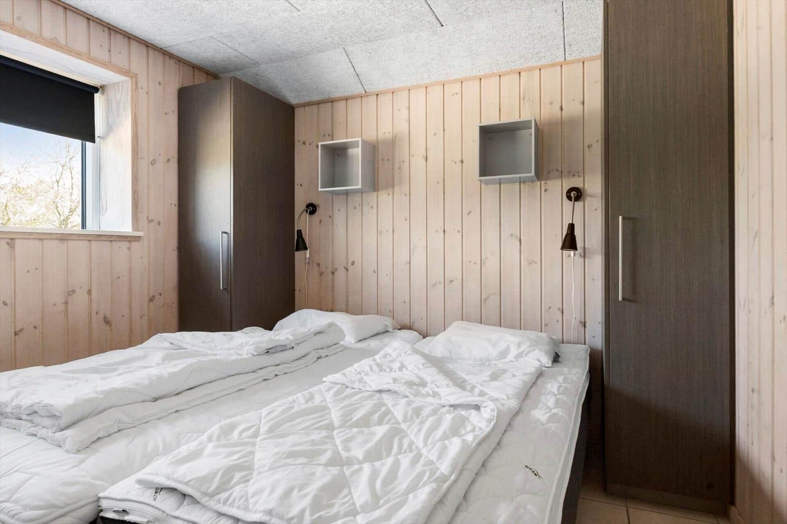 Schlafzimmer mit Holzwand, Bett und Schränken. Fenster mit Blick auf Bäume.
