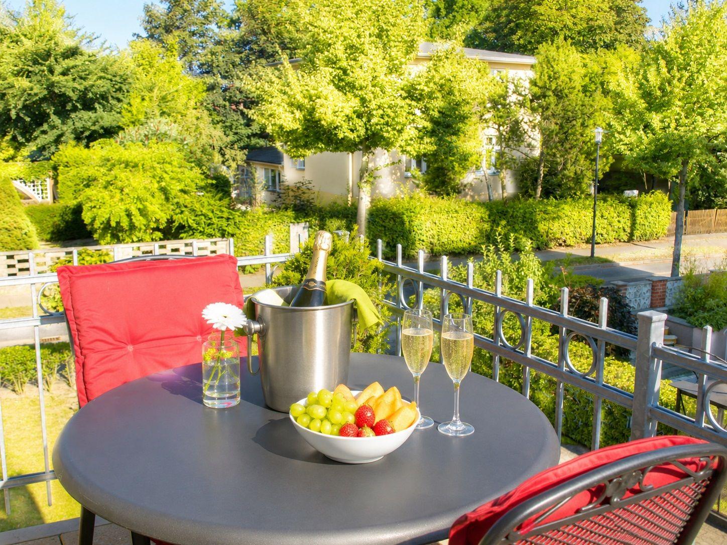 Auf der Terrasse steht ein Tisch mit Champagner, Obst und Stühlen mit roten Kissen.