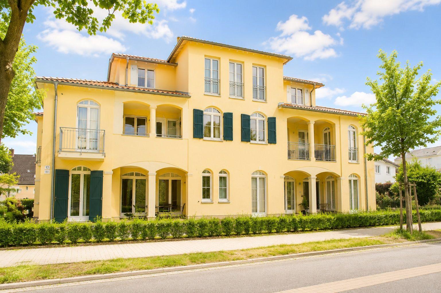 Gelbes Haus mit Balkonen und grüner Hecke an der Straße.