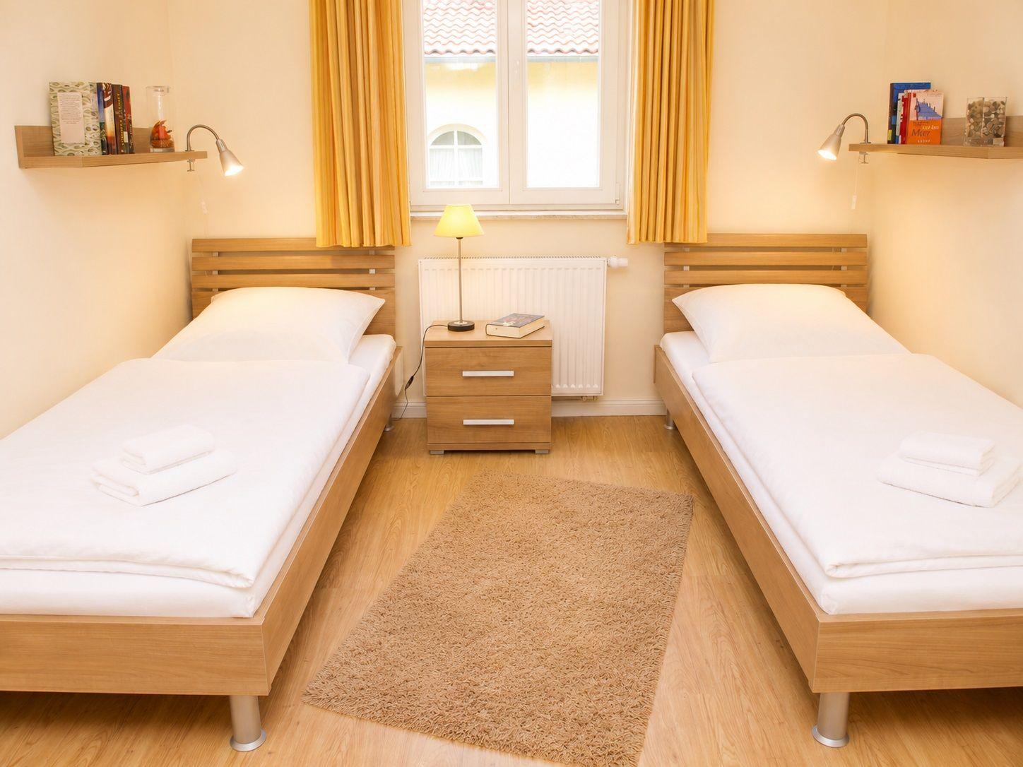 Zwei Einzelbetten mit weißen Laken, Nachttisch und Fenster mit gelben Vorhängen.
