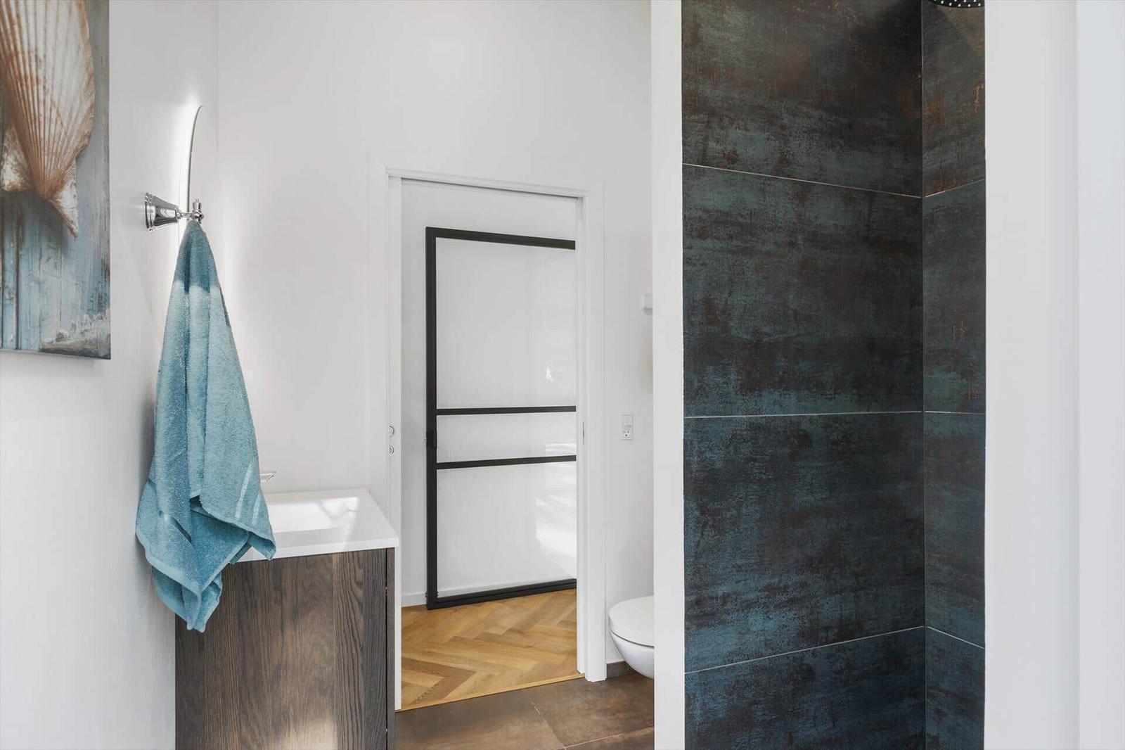 Badezimmer mit Duschbereich, Waschbecken und Toilette. Wand mit dunklen Fliesen.