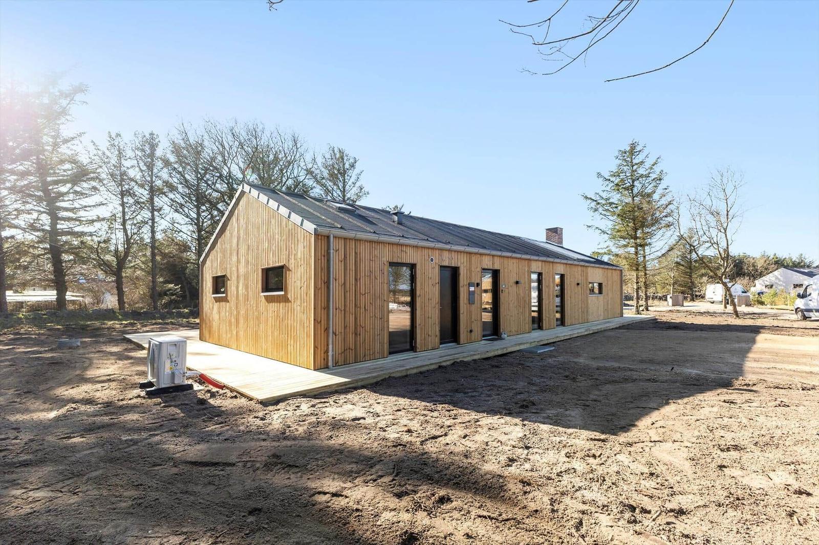 Moderne Holzhütte mit mehreren Eingängen und Fenstern, umgeben von Bäumen und freiem Gelände.