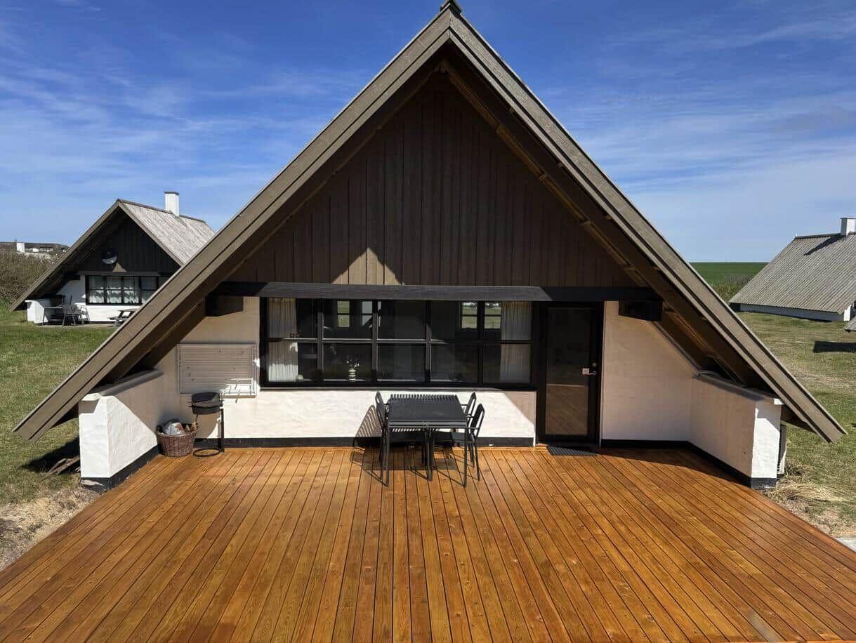 Haus mit Holzterrasse, Sitzgruppe und Grill. Blick auf grüne Wiesen.