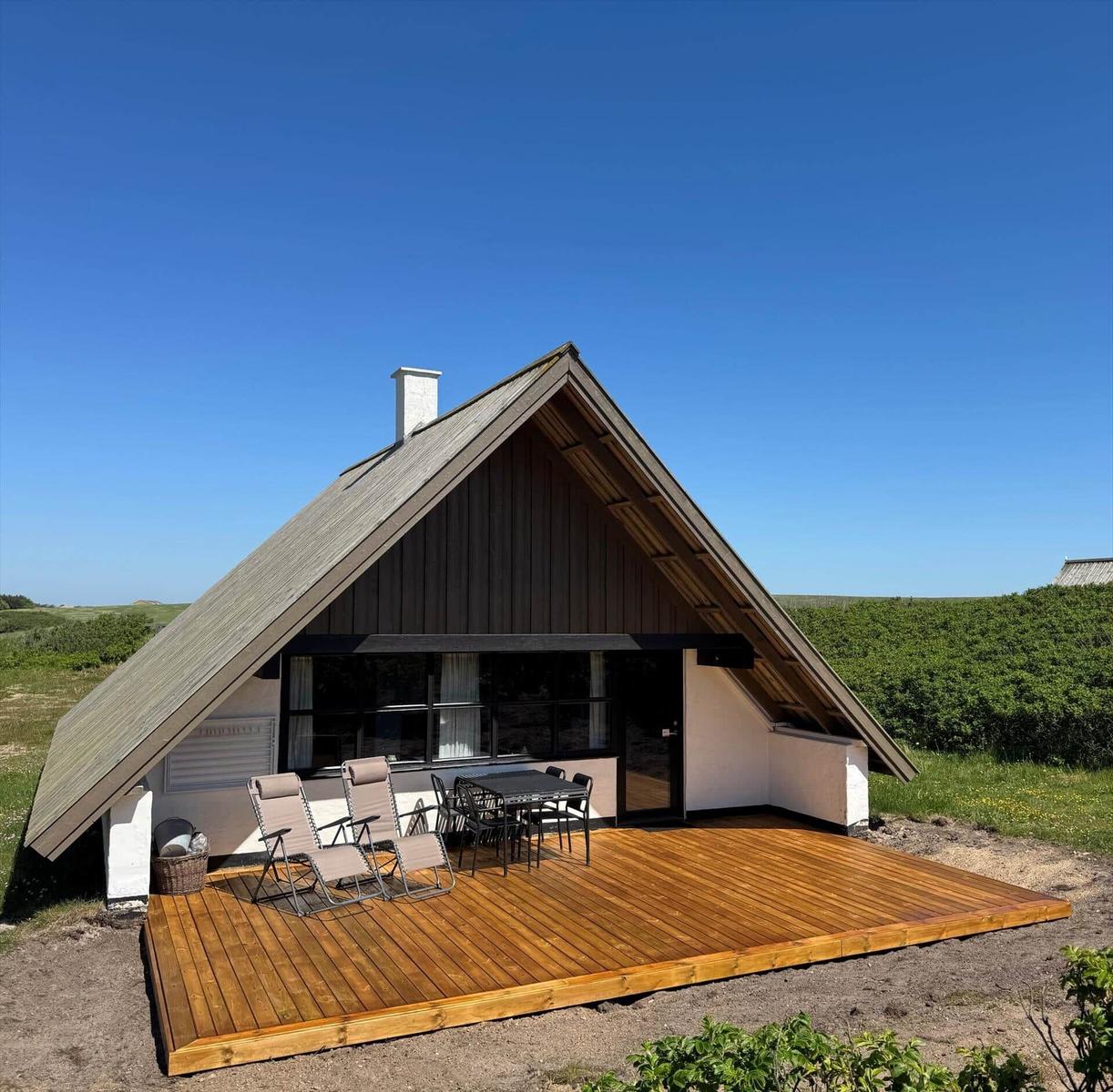 A-frame-Haus mit Holzterrasse, Liegestühlen und Tisch unter blauem Himmel.