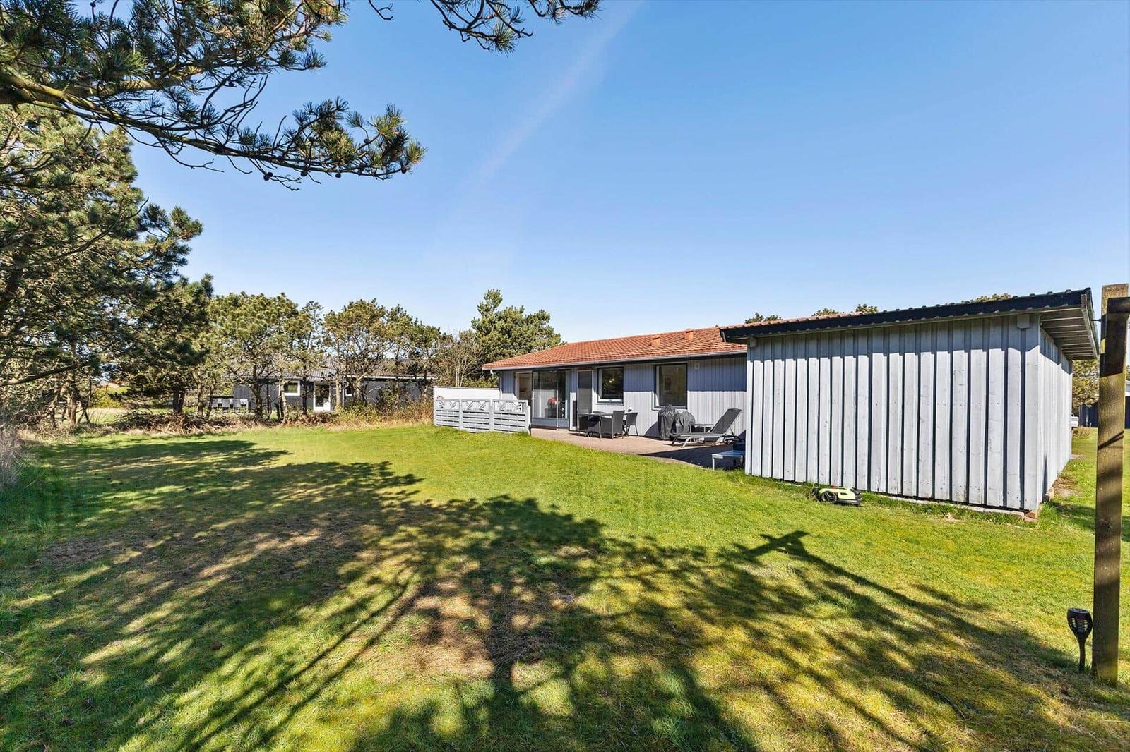 Ein Ferienhaus mit Terrasse, Garten und Bäumen unter blauem Himmel.