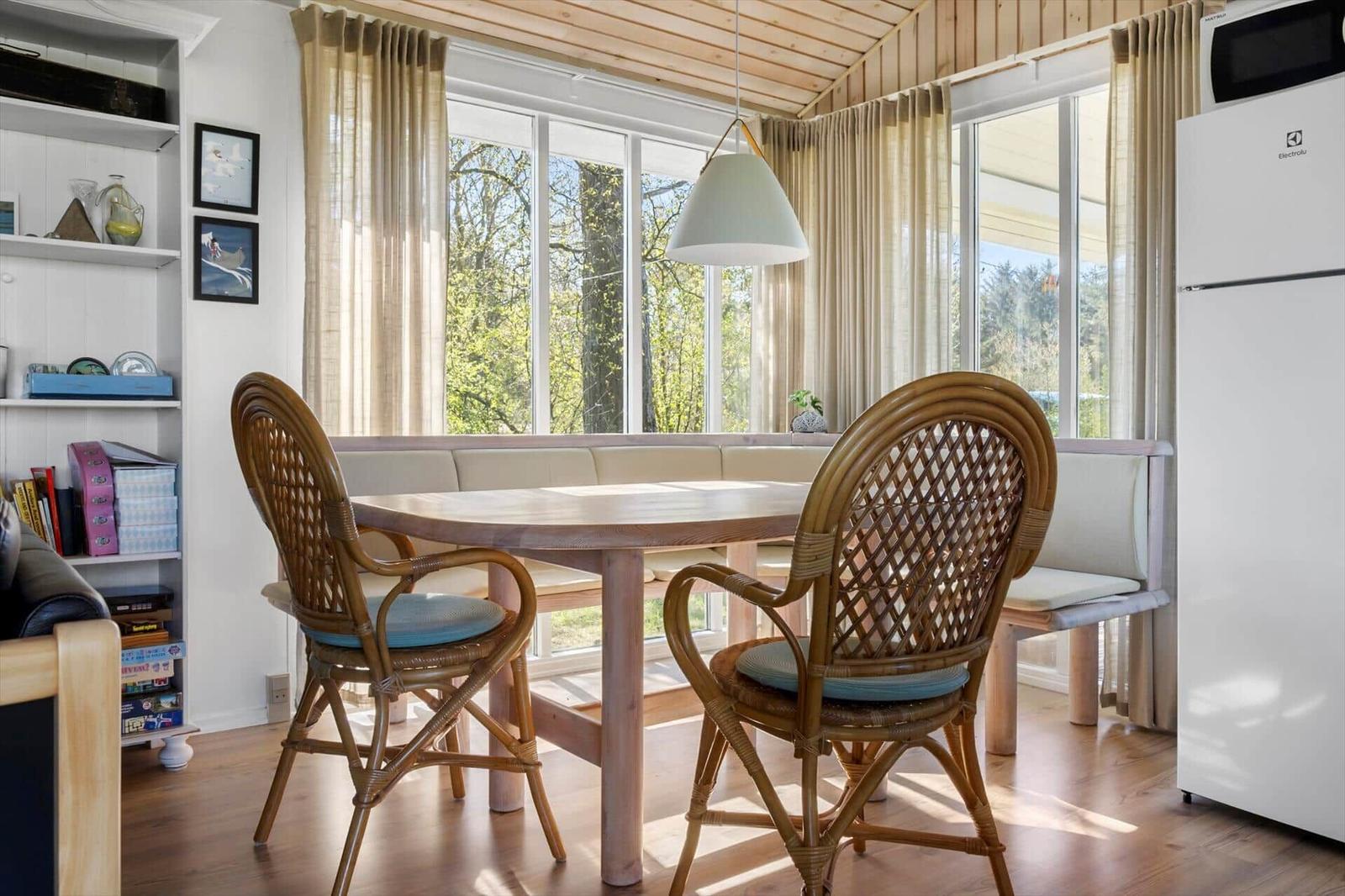 Dining area with wooden table, rattan chairs, and bench. Large windows, refrigerator, and bookshelf.