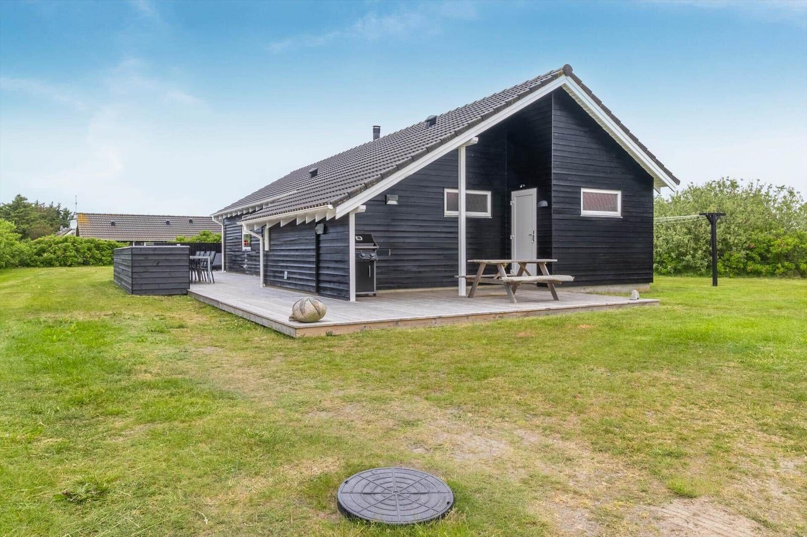 Schwarzes Holzhaus mit Terrasse, Grill und Picknicktisch auf grünem Grund.