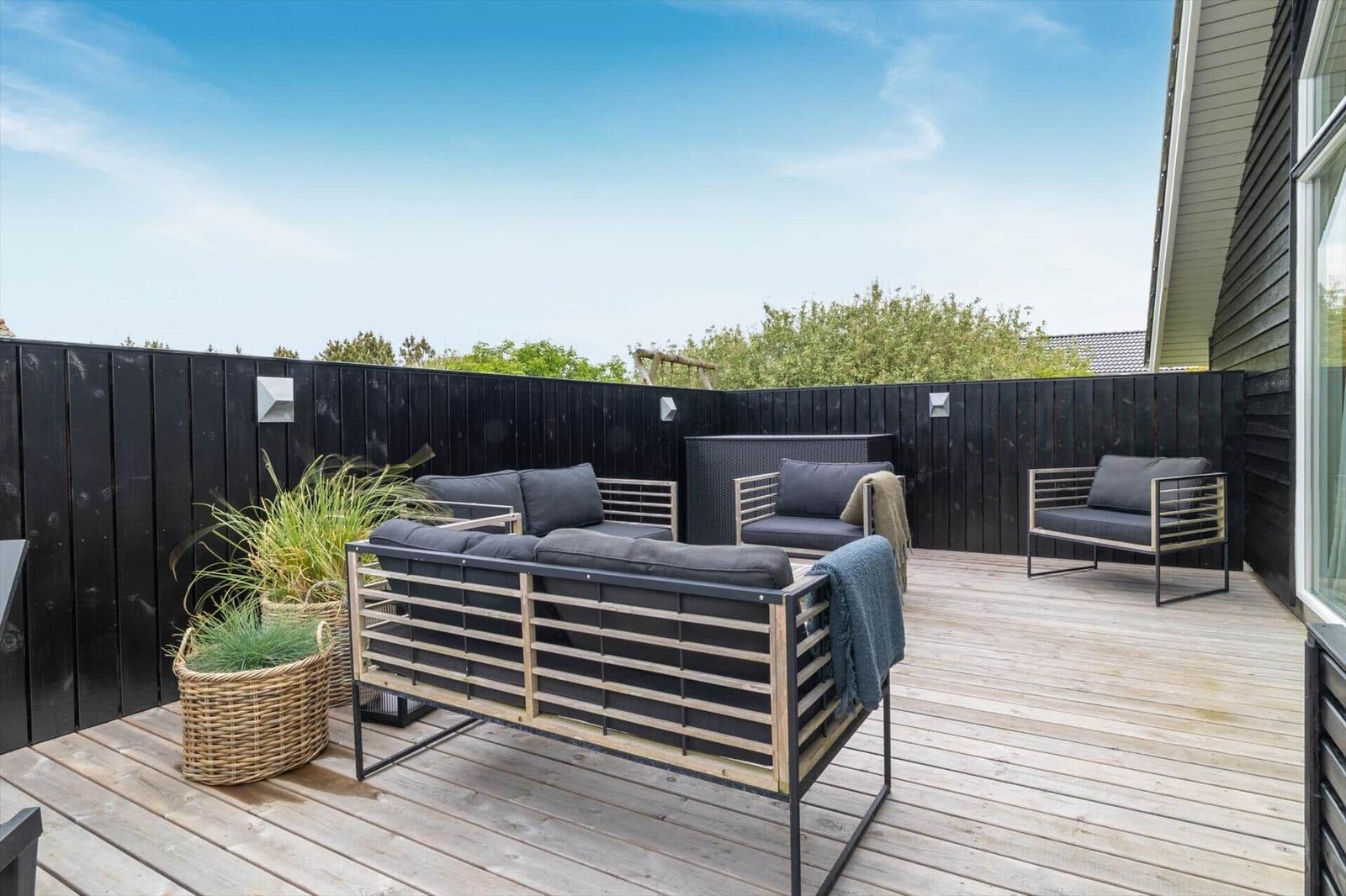 Terrasse mit Sitzgruppe, Pflanzen und Holzdielen. Schwarze Umzäunung und Himmel.