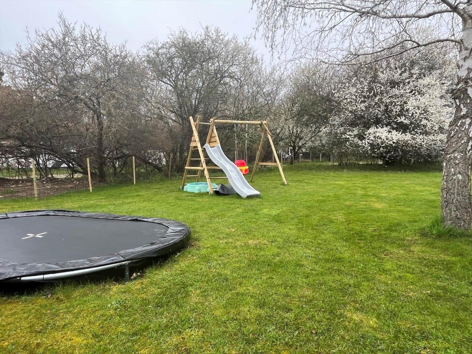 Large garden with trampoline, slide, and swing. Trees with blossoms in the background.
