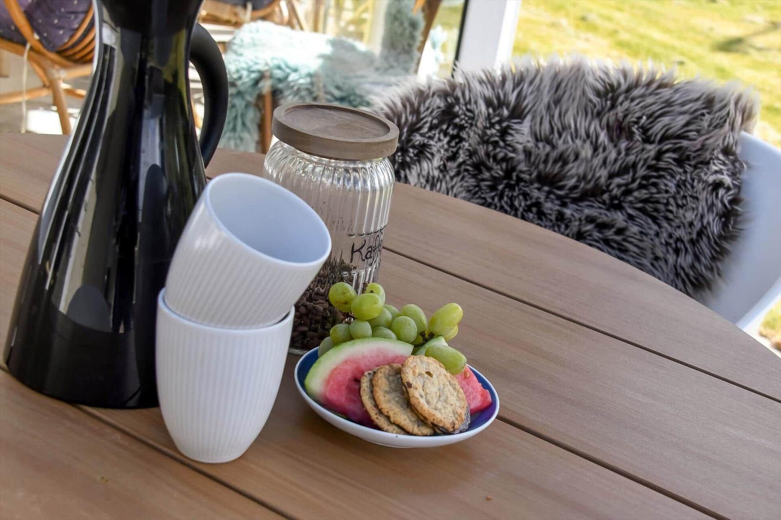 Holztisch mit Kaffeekanne, Tassen, Obst und Keksen. Hintergrund: Sitzgelegenheit mit Felldecke.