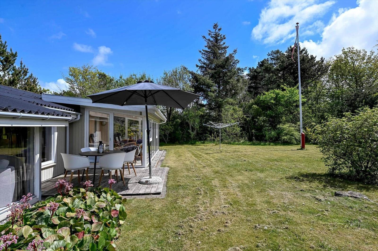Haus mit Terrasse, Sonnenschirm und Garten. Hintergrund: Bäume und klarer Himmel.