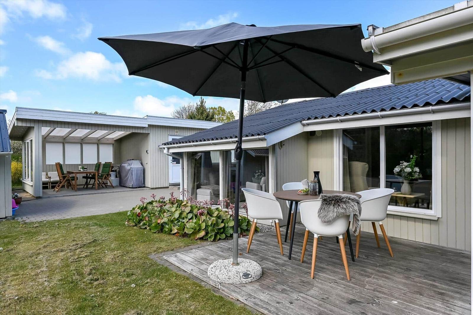Terrasse mit Tisch, Stühlen und Sonnenschirm. Hintergrund: Haus mit Glasfront und Garten.