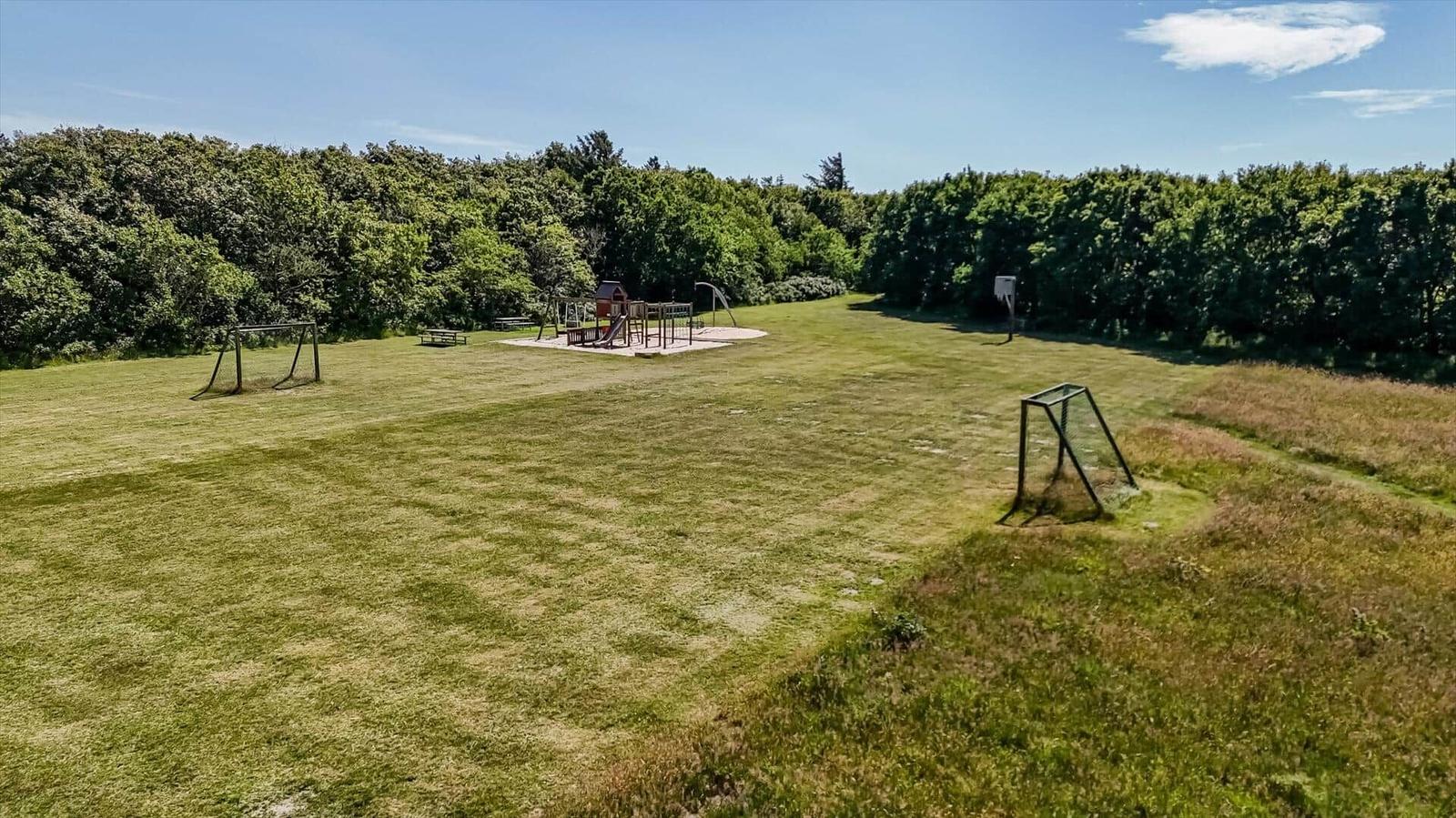 Großes Grünfeld mit Spielplatz und Fußballtoren. Umgeben von Bäumen unter blauem Himmel.
