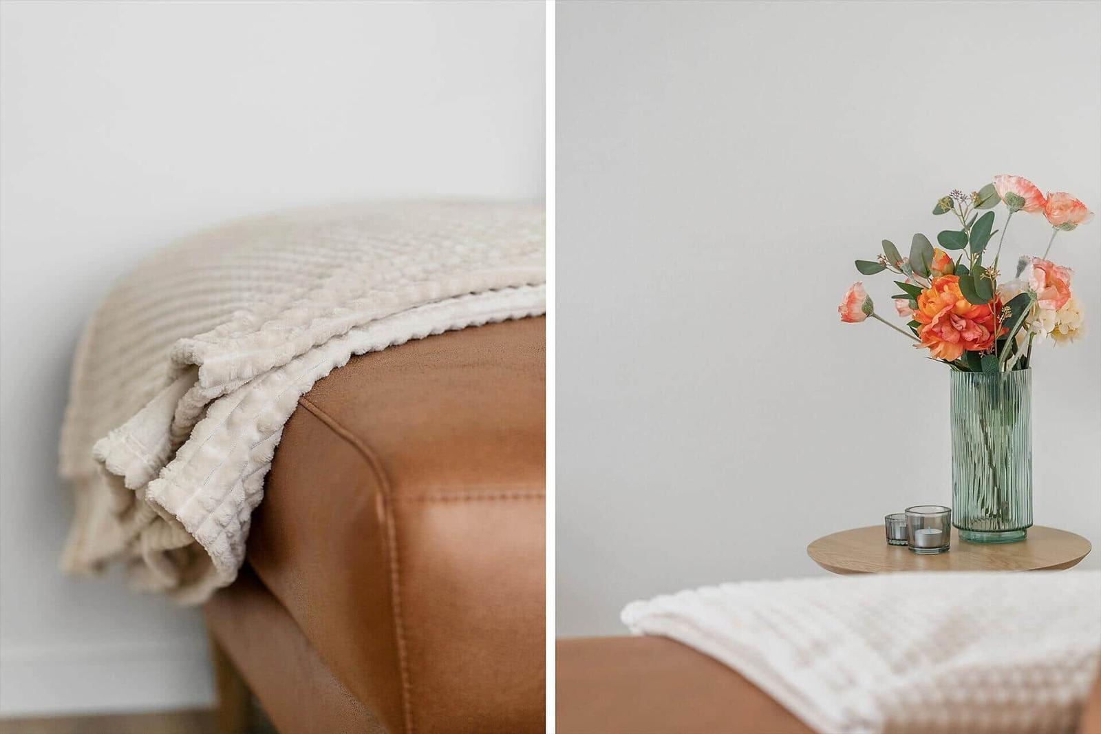 Beige blanket on brown leather sofa. Flowers in glass vase on wooden table.