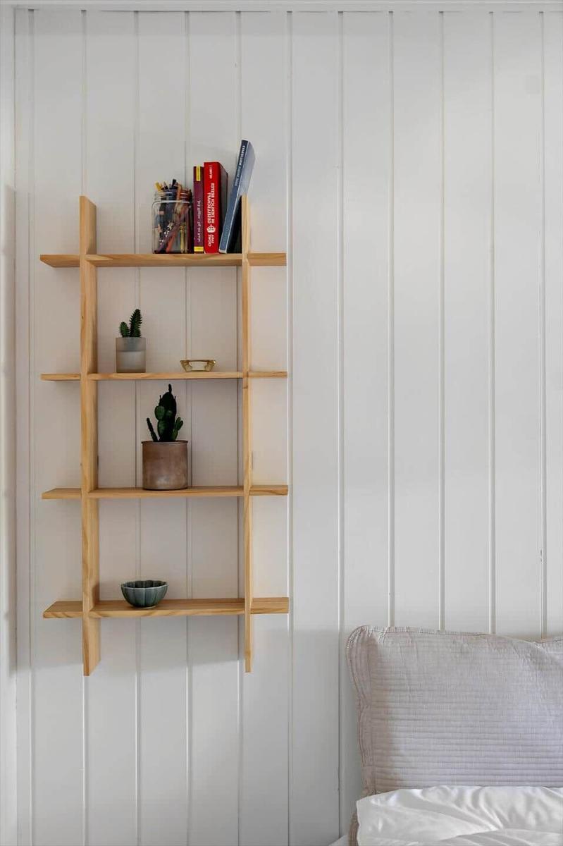 Holzregal an weißer Wand mit Büchern, Pflanzen und Schale. Bett mit Kissen rechts.