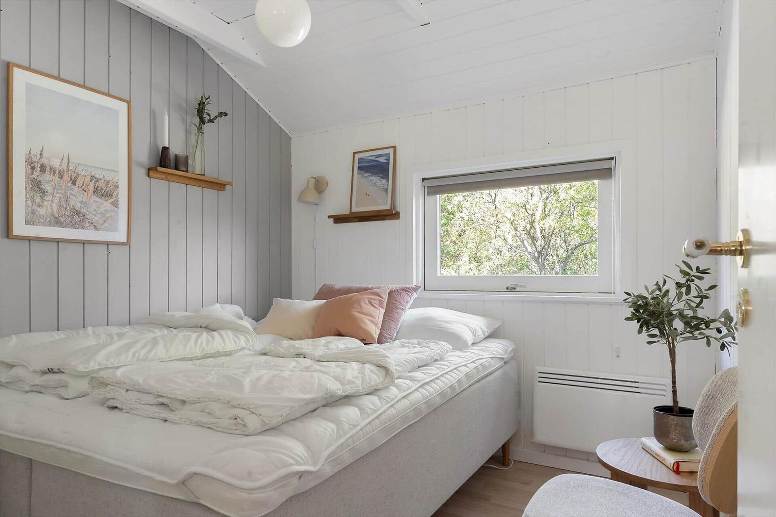 Schlafzimmer mit Bett, Fenster und Pflanze. Wände sind weiß und grau verkleidet.