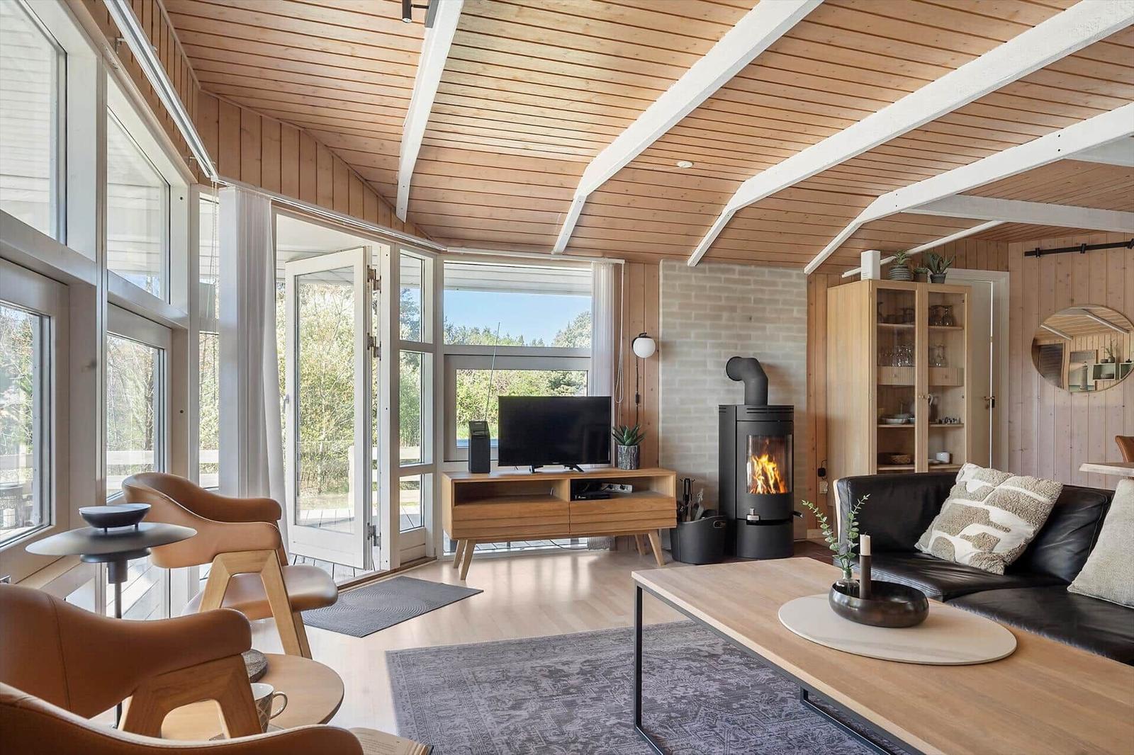 Wohnzimmer mit Holzdecke, Kamin und großem Fenster. Möbel sind modern und schlicht.