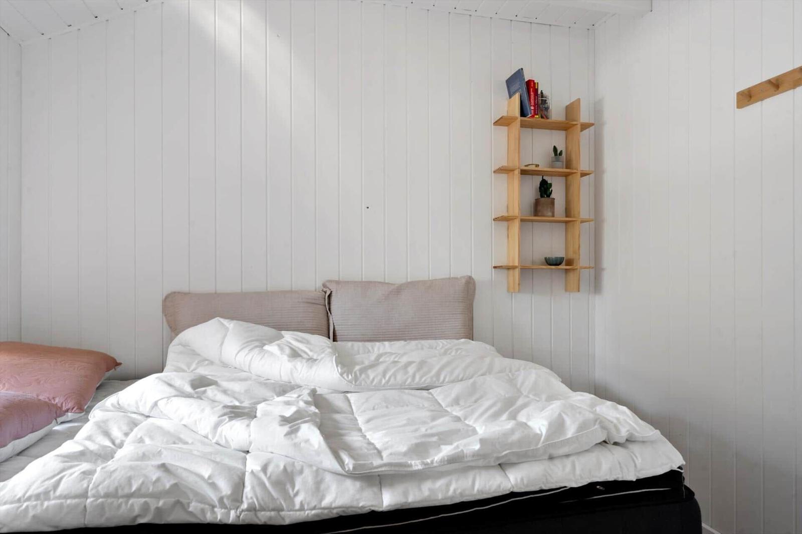 Ein Bett mit weißer Bettwäsche und rosa Kissen. An der Wand hängt ein Holzregal mit Büchern und Pflanzen.