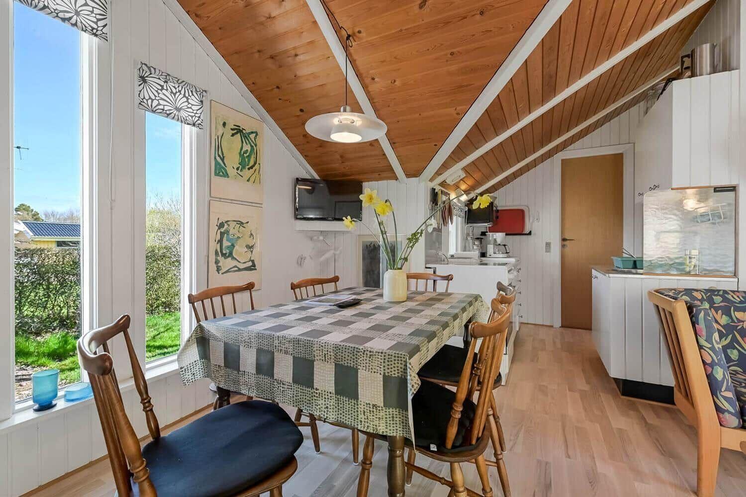 Esszimmer mit Holztisch, Stühlen und Küchenbereich. Große Fenster mit Blick auf Garten.