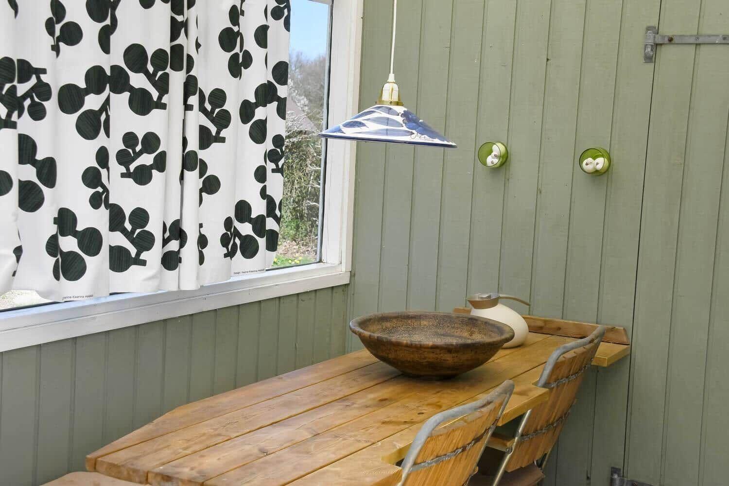 Holztisch mit Schale und Stühlen unter einer Lampe neben Fenster mit Vorhang.