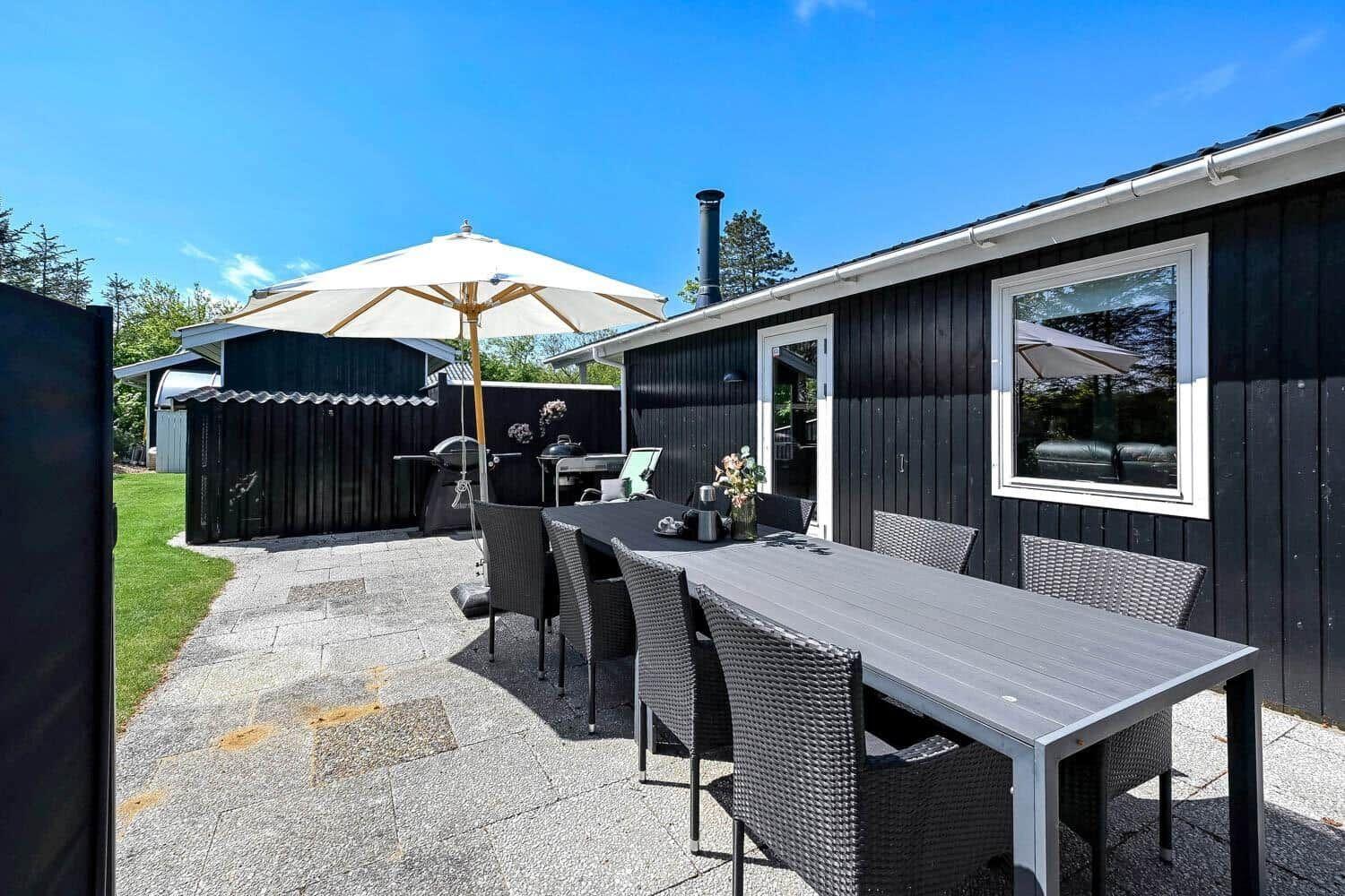 Terrace with table, chairs, and umbrella. Background: black-painted house with window.