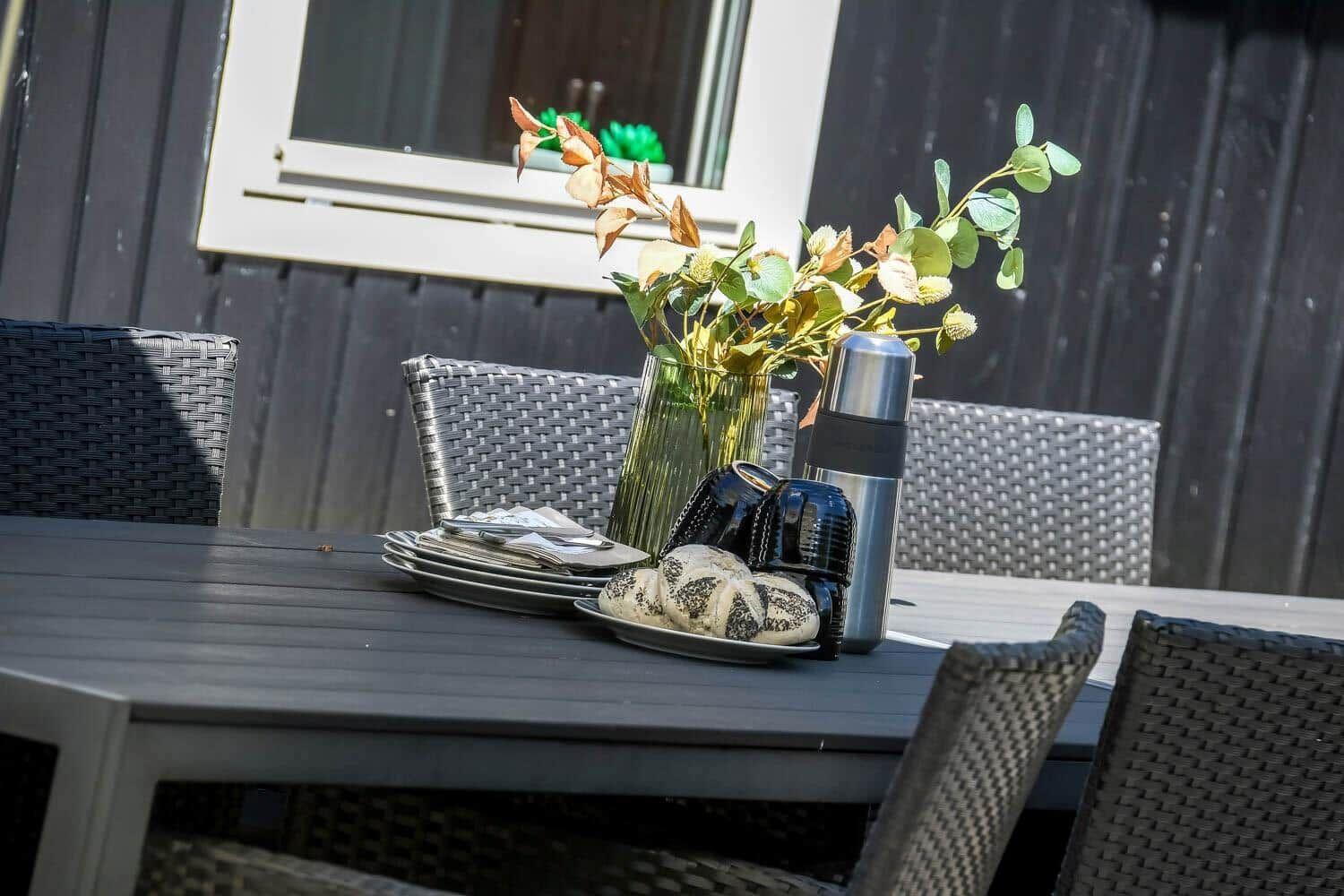 Outdoor area with table, chairs, and floral arrangement. On the table are plates, mugs, and a thermos.