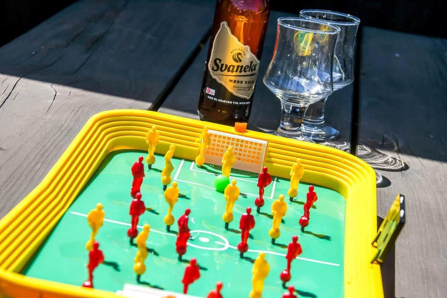Foosball table with red and yellow figures, bottle and two glasses on wooden table.