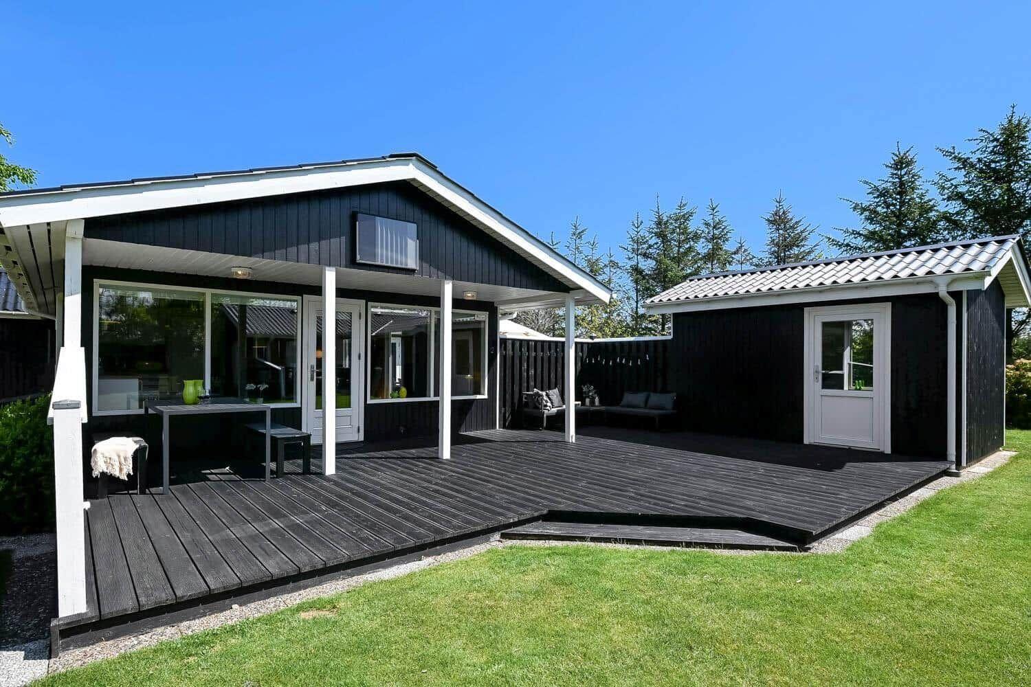 Black wooden house with terrace and garden. White windows and door. Background with trees.