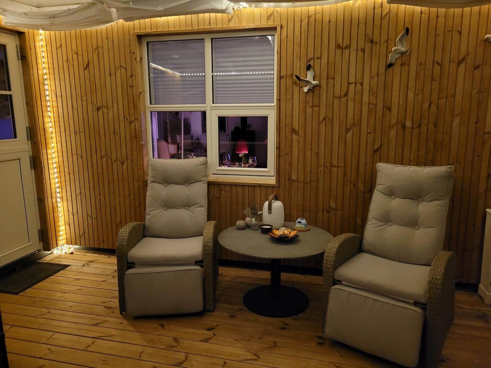 Zwei bequeme Sessel stehen um einen runden Tisch herum. Holzwände und Boden. Fenster mit Jalousien.