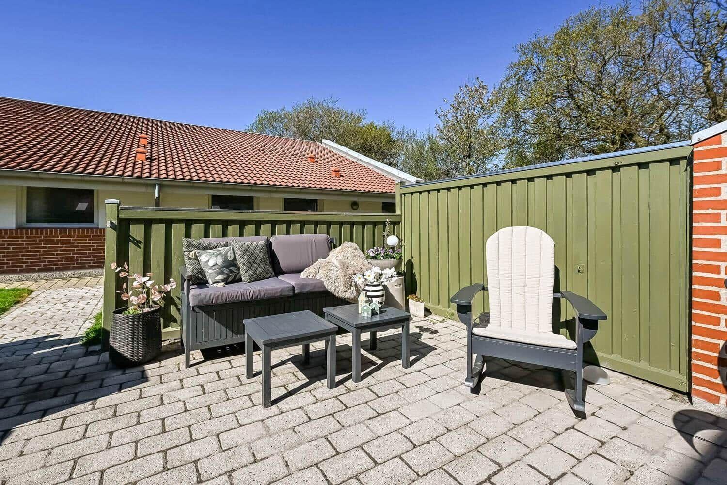 Terrace with seating group, rocking chair, and plants. Background: house with red roof and green fence.