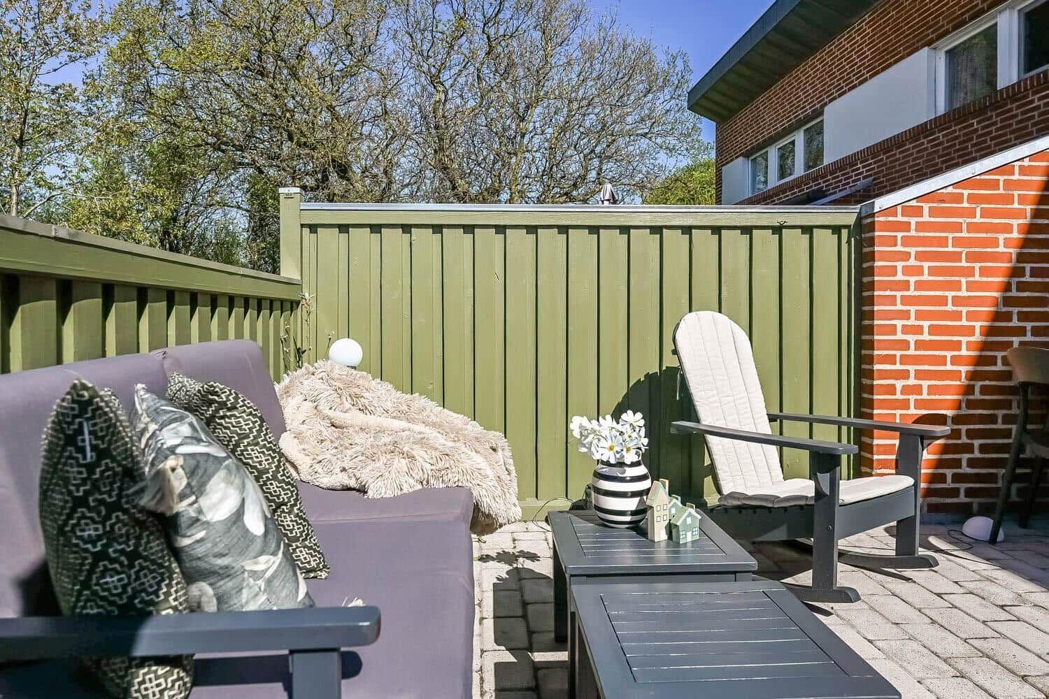 Terrace with sofa, chair, and table. Background: green fence and brick house.