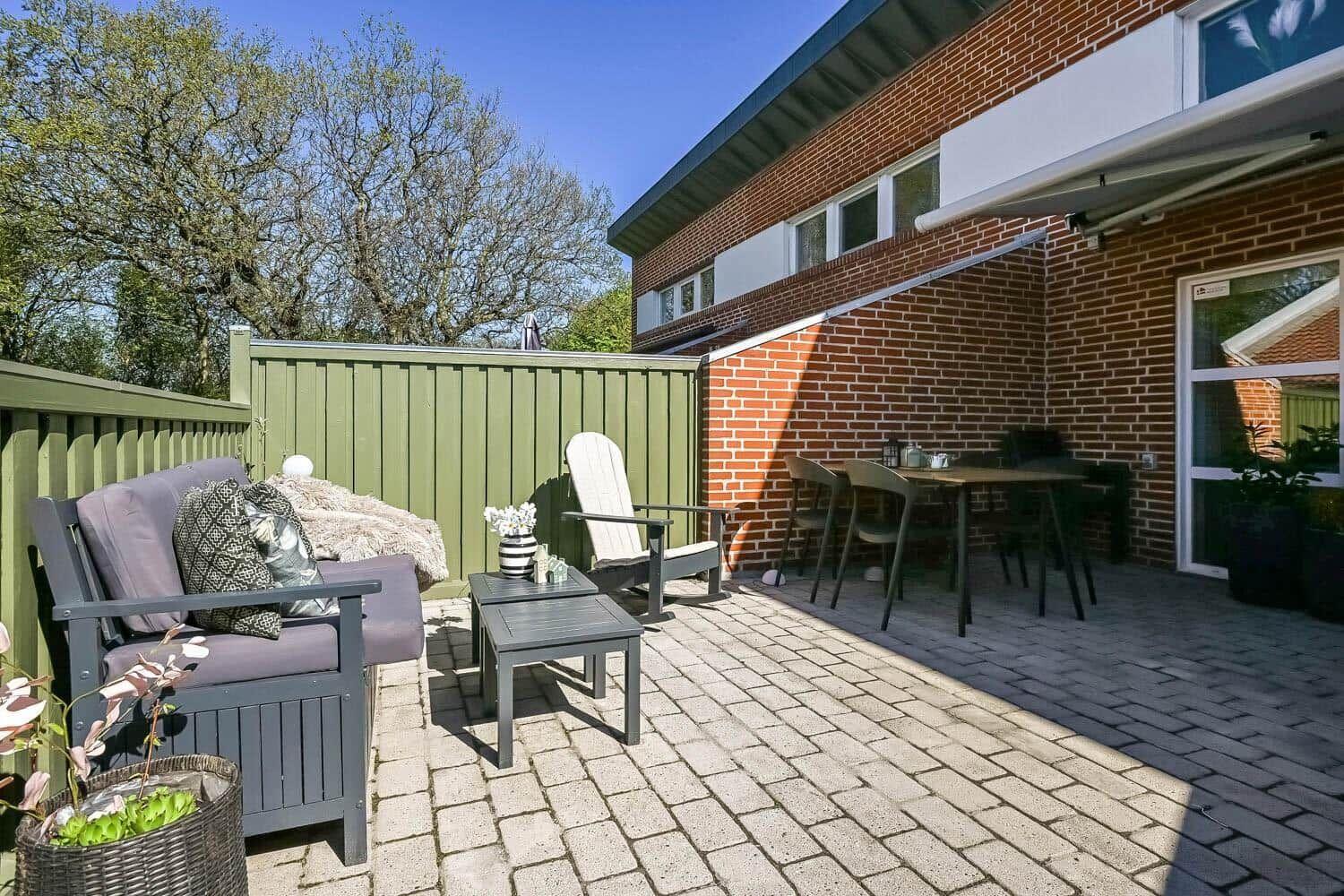 Terrace with seating group, table and chairs. Background: green fence and brick house.