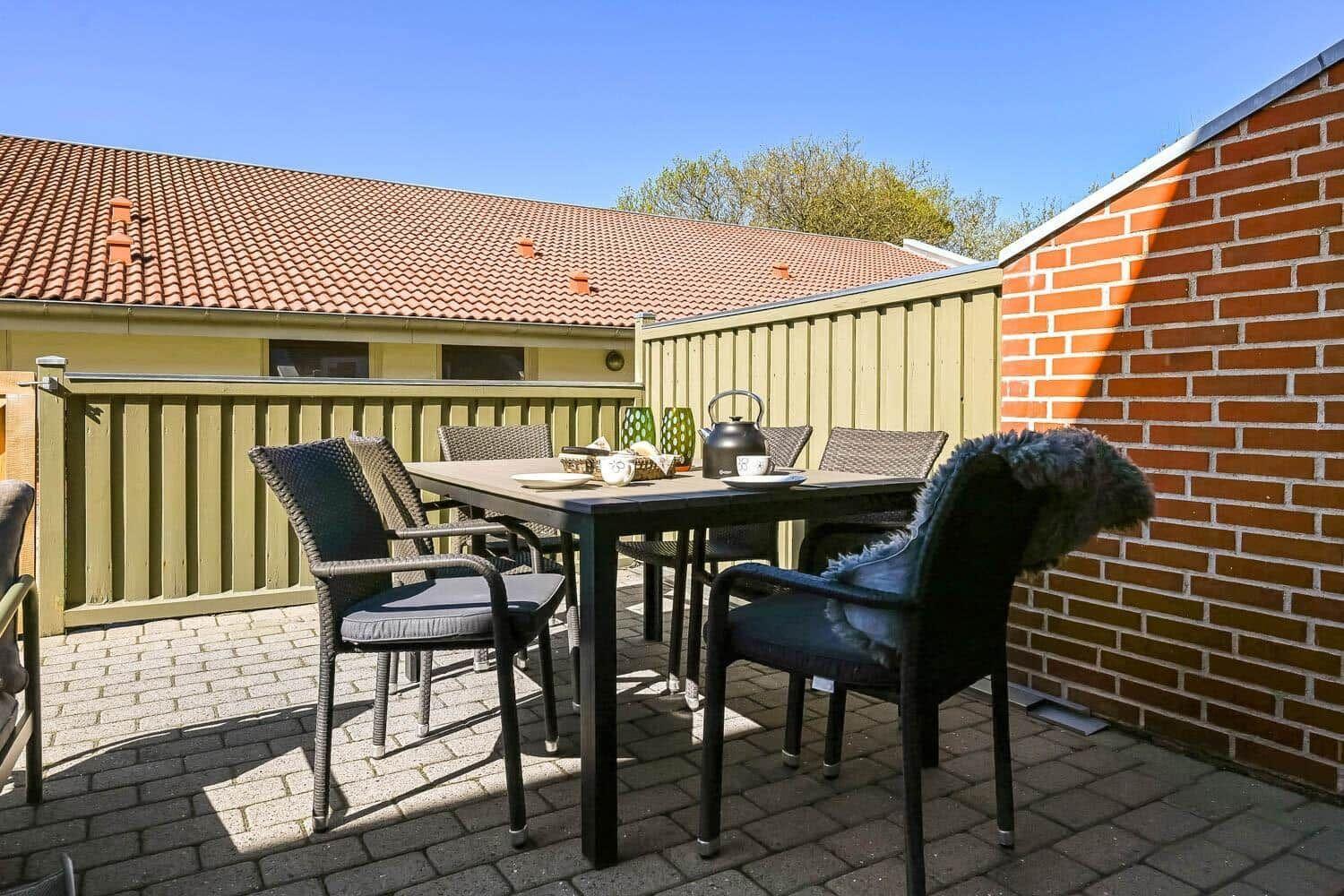 Terrasse mit Tisch, Stühlen und Teekanne. Hintergrund: Haus mit Ziegeldach und Holzgeländer.