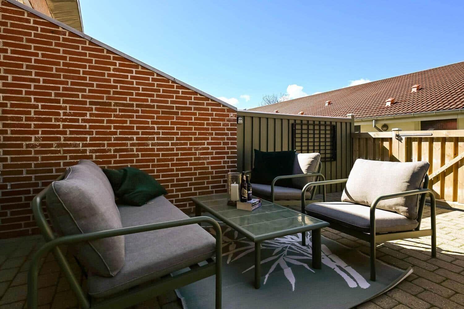 Terrasse mit Sitzgruppe, Tisch und Teppich. Hintergrund: Backsteinwand und Holzzaun.