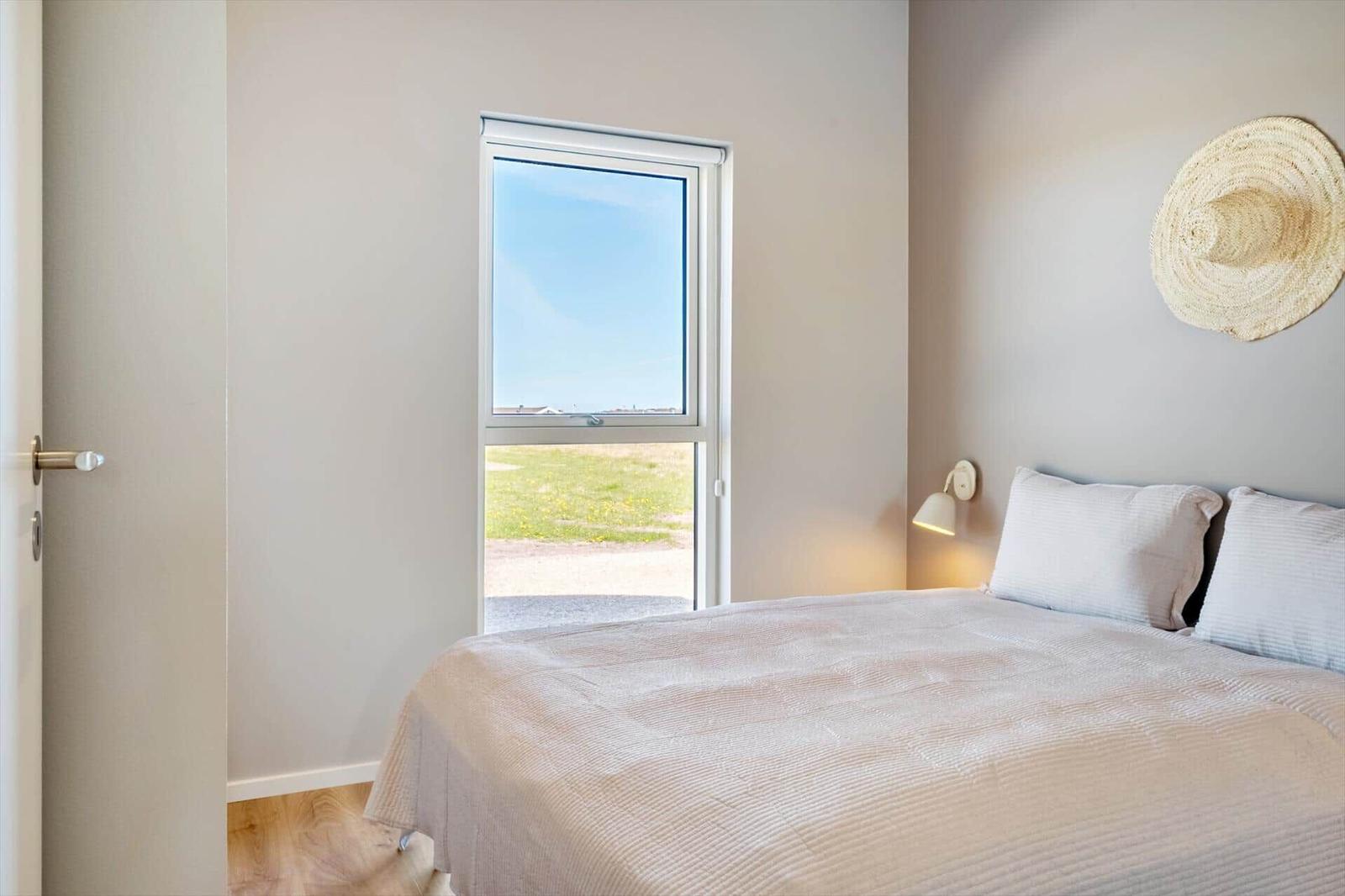 Schlafzimmer mit Bett, Fenster und Wandlampe. An der Wand hängt ein Strohhut.