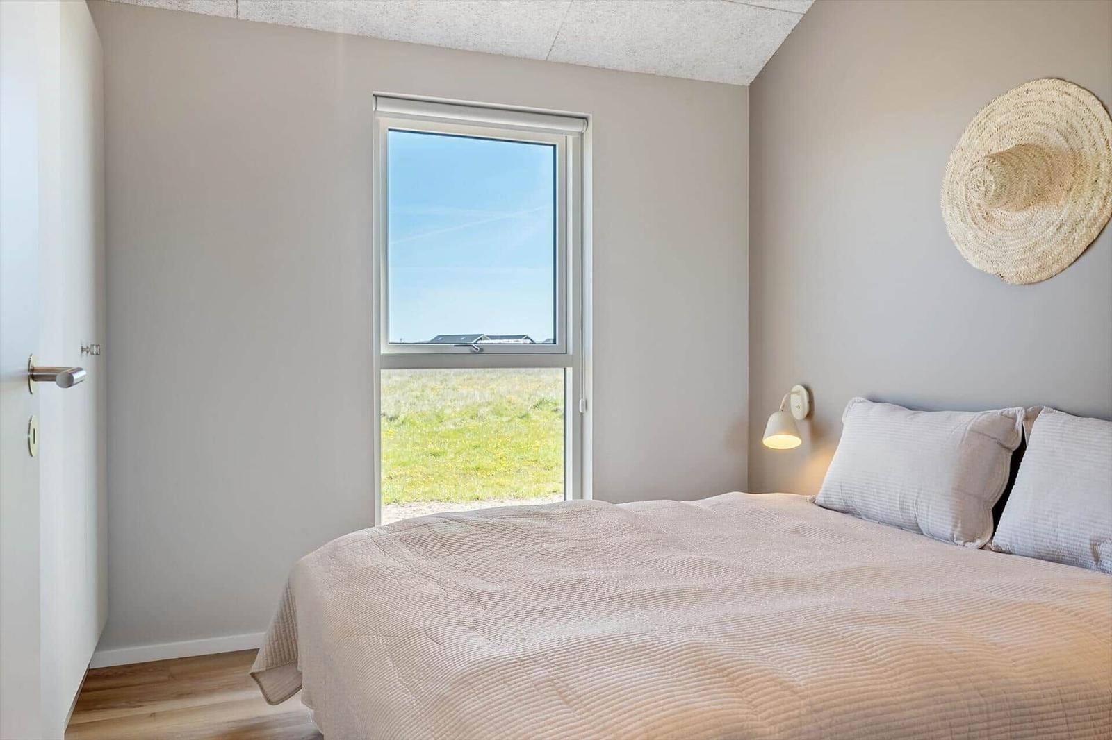 Schlafzimmer mit Bett, Fenster und Wandlampe. Draußen ist Gras und Himmel sichtbar.