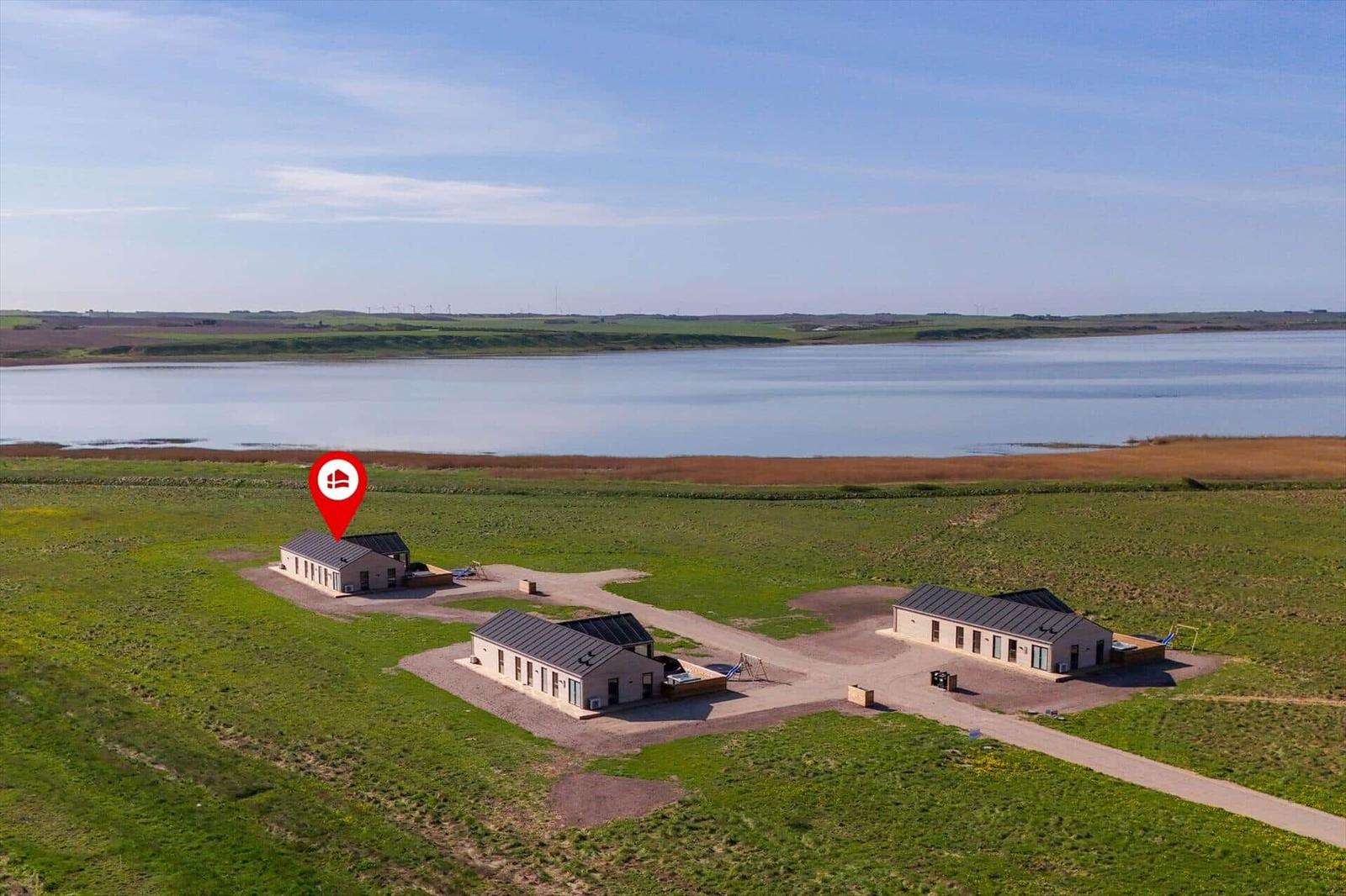 Drei Häuser mit Dach und Terrasse stehen auf einer grünen Wiese neben einem See.