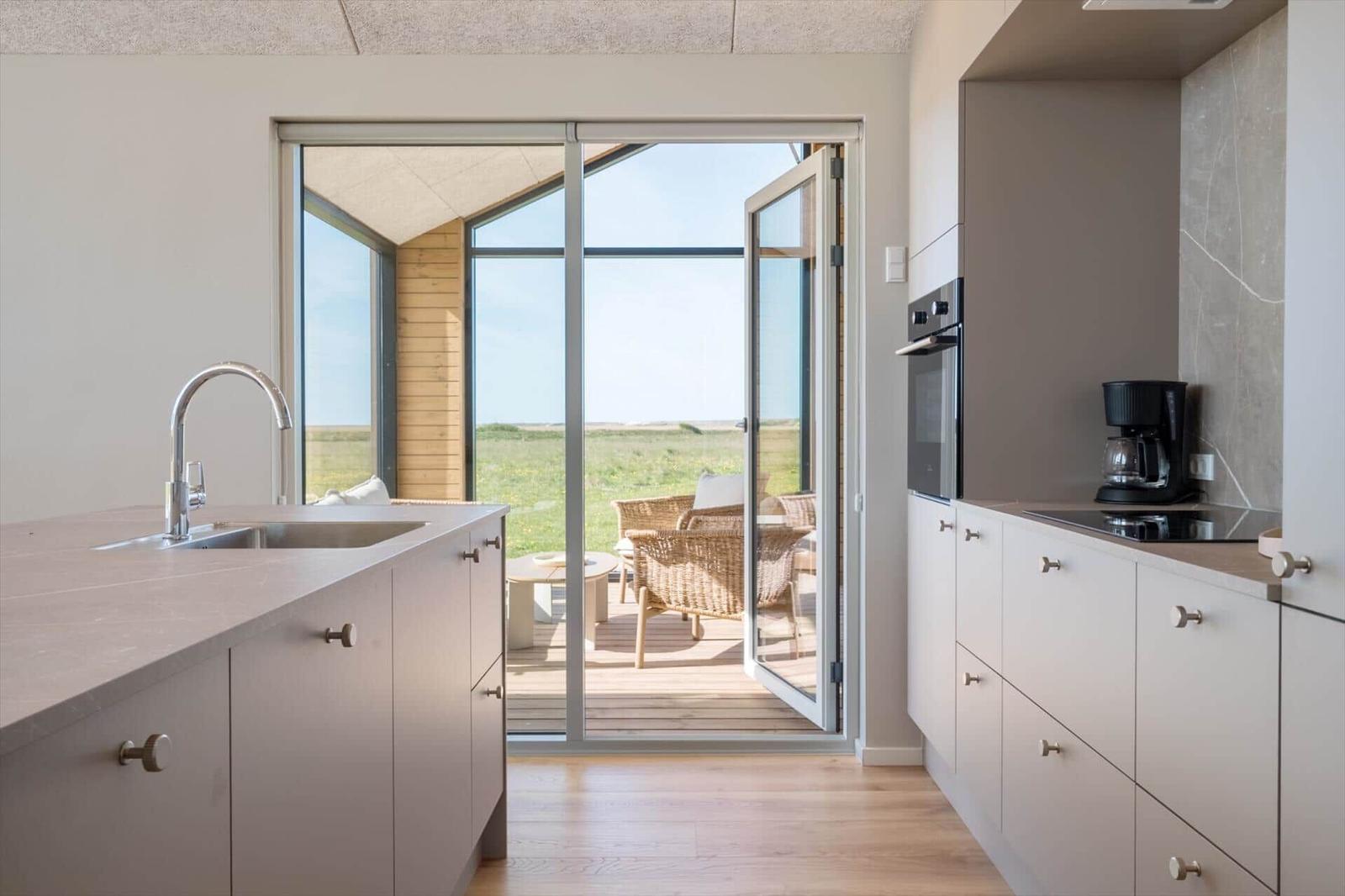 Küche mit Blick auf Terrasse und Grasland. Glastüren, Holzboden, Waschbecken, Kaffeemaschine.