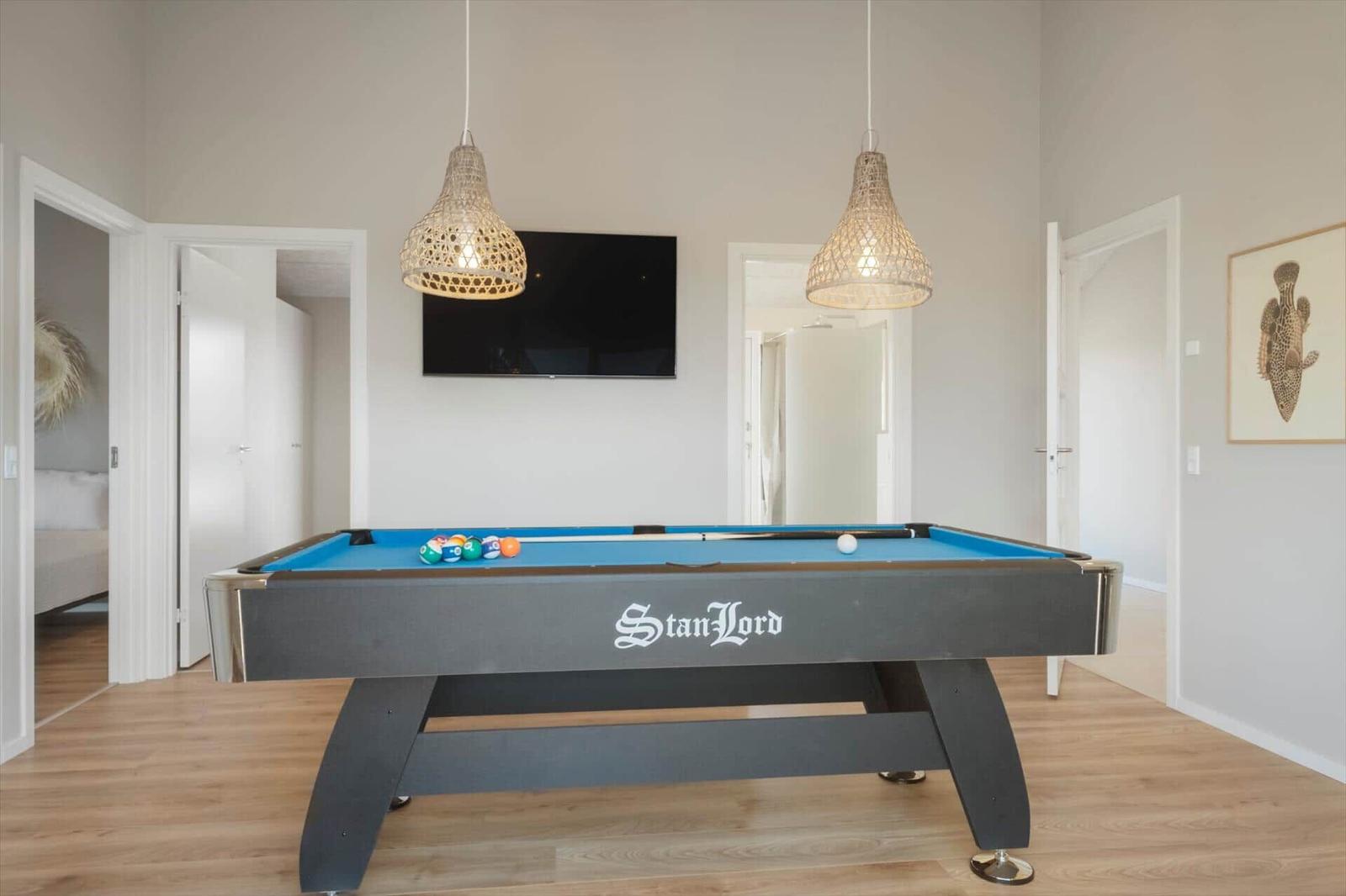 Schwarzer Billardtisch mit blauem Tuch und Kugeln. Zwei Hängelampen und Fernseher an der Wand.
